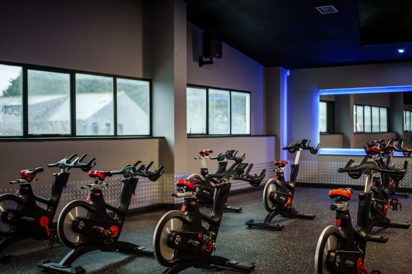 Indoor cycling studio with multiple stationary bicycles arranged in rows, illuminated by soft indoor lighting, with large windows on the side and a ceiling-mounted TV or screen.