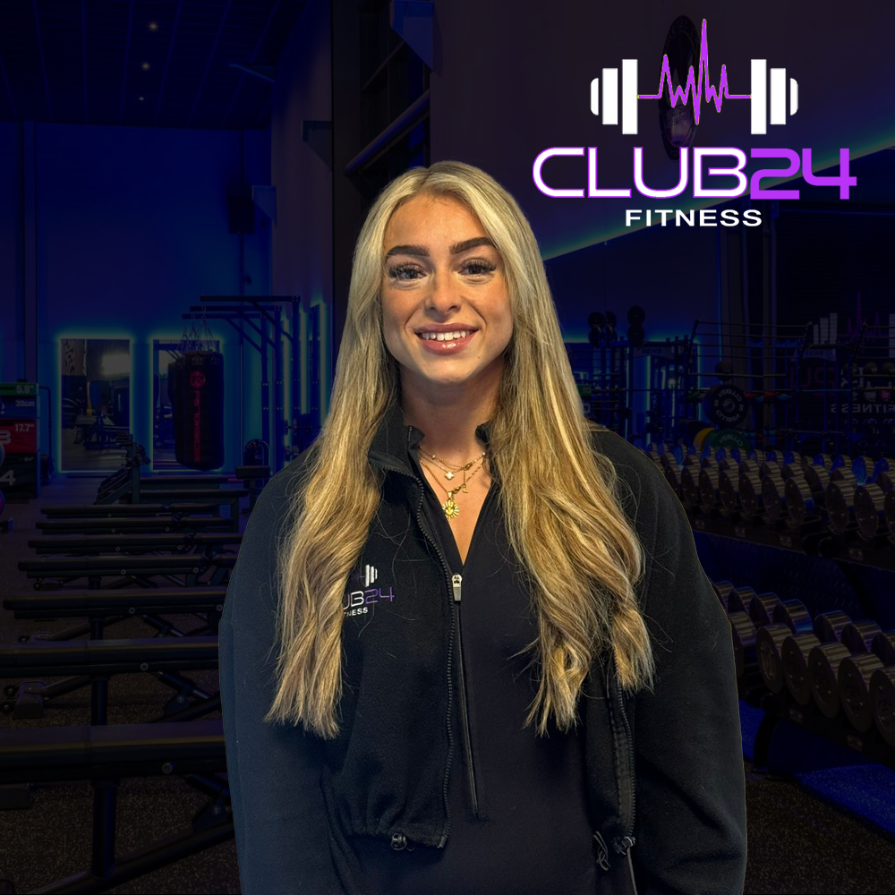 A woman with long blonde hair smiling inside a gym. She is wearing a black zip-up jacket with the logo 'CLUB24 FITNESS' on it. The background features gym equipment and a sign with the same logo.