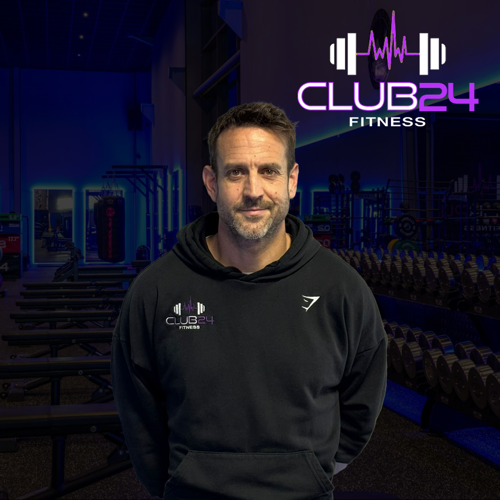 Man in a black hoodie with a 'Club 24 Fitness' logo standing inside a gym with fitness equipment and a neon sign with the same logo in the background.