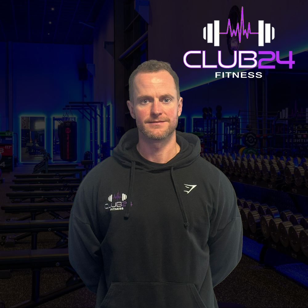 Man standing in a gym with rows of dumbbells, yoga mats, and workout equipment; logo for Club 24 Fitness displayed in the background.