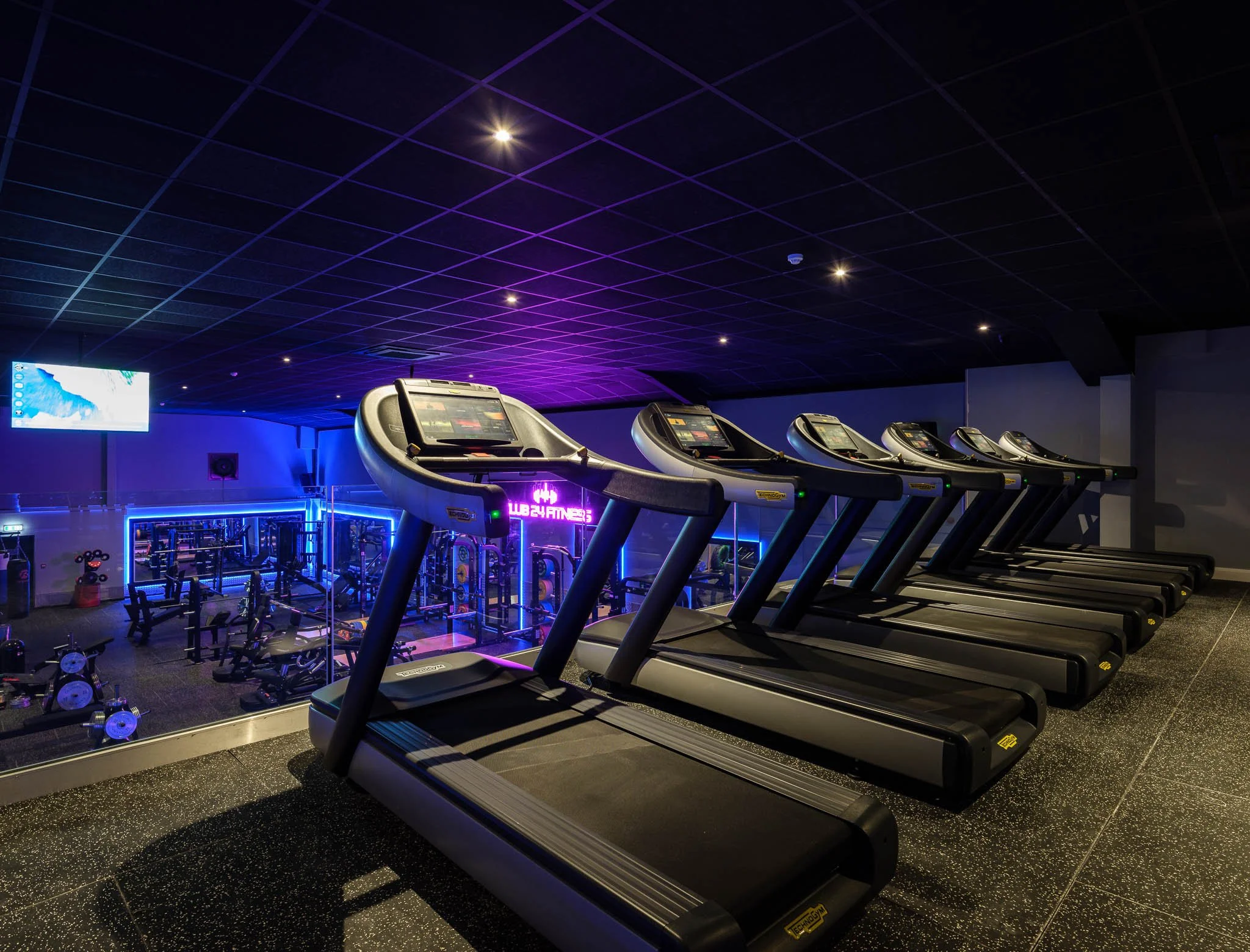 Row of six treadmills in a modern gym with neon lights and fitness equipment in the background