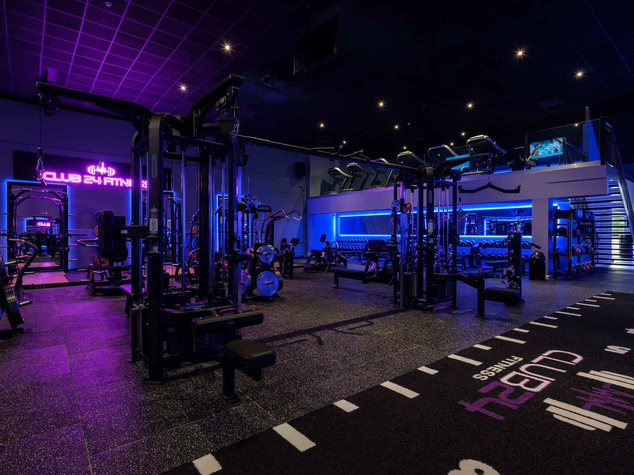 A neon-lit gym with black equipment, illuminated purple and blue walls, a staircase, and a branded mat reading 'Club 24 Fitness' with purple and white markings.
