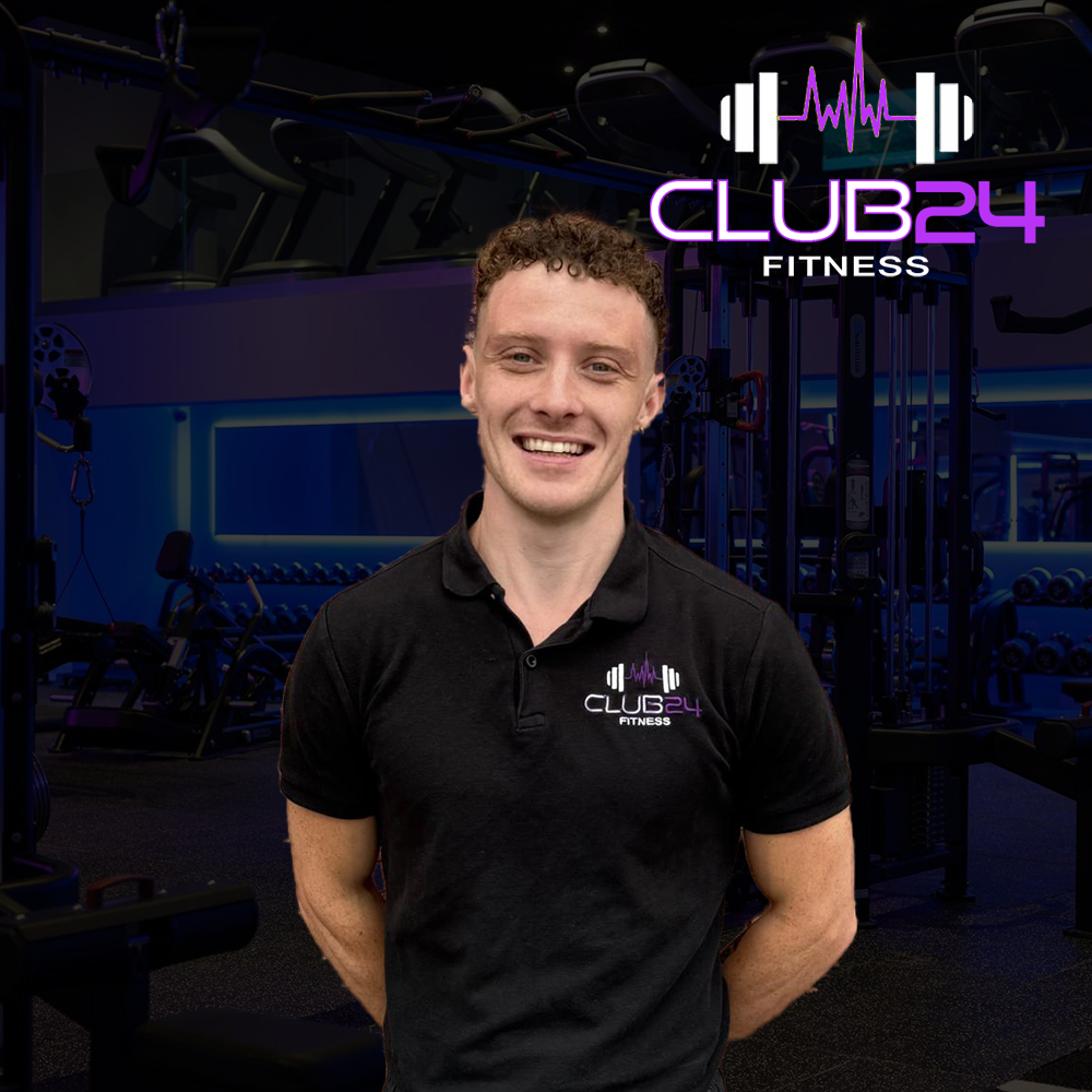 A smiling man in a black polo shirt with the Club24 Fitness logo stands in a gym, with workout equipment and a neon sign in the background.