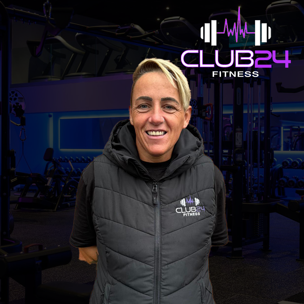 A smiling woman in a black vest with 'Club 24 Fitness' logo standing inside a gym with exercise equipment and a neon sign.