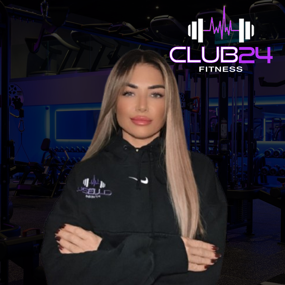 A woman with long blonde hair standing in a gym, wearing a black hoodie with the logo 'Club24' on it. The gym has exercise equipment and a sign that reads 'Club24 Fitness' with a barbell and sound wave graphic in the background.