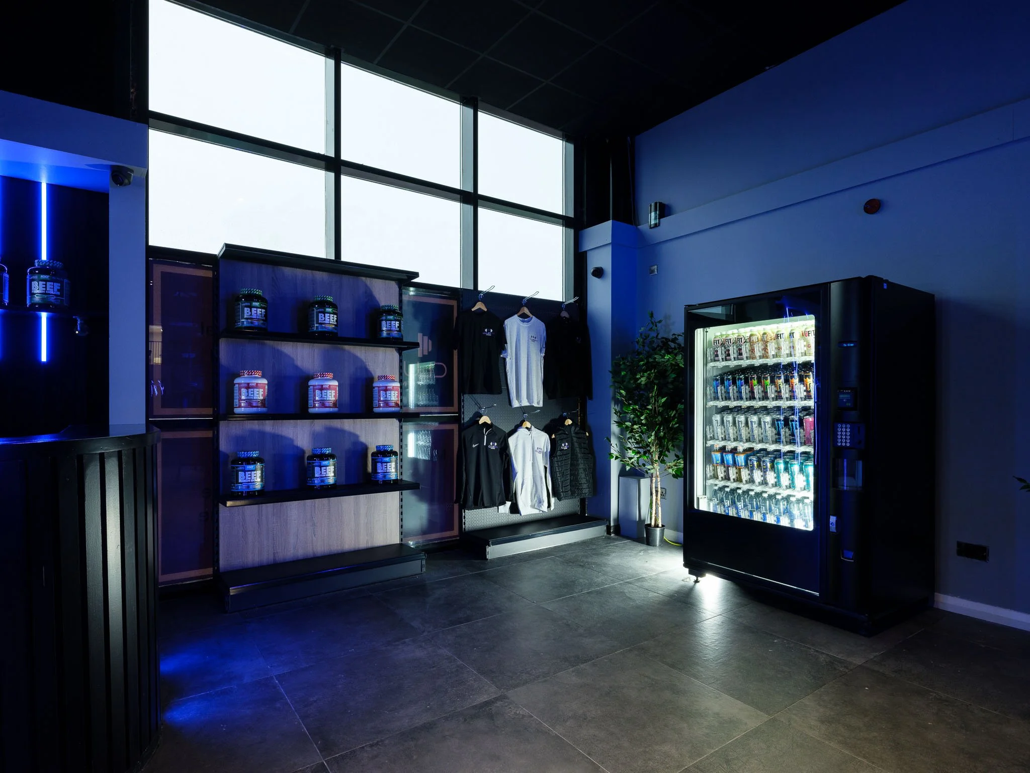 Interior of a retail store with large windows, display shelves, hanging clothing, a vending machine, and a potted plant.