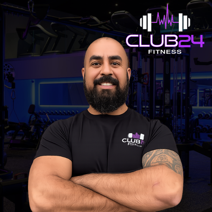 A man with a beard and tattooed arm standing with arms crossed inside a gym, with the Club24 Fitness logo and a purple workout icon in the background.