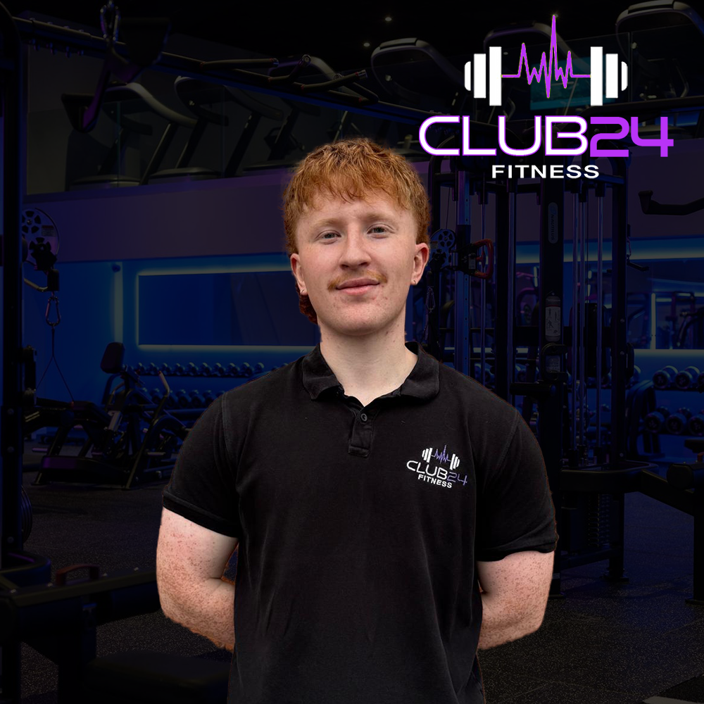 A man in a black polo shirt with a 'Club 24 Fitness' logo stands inside a gym with workout equipment and a neon sign with the gym's logo in the background.
