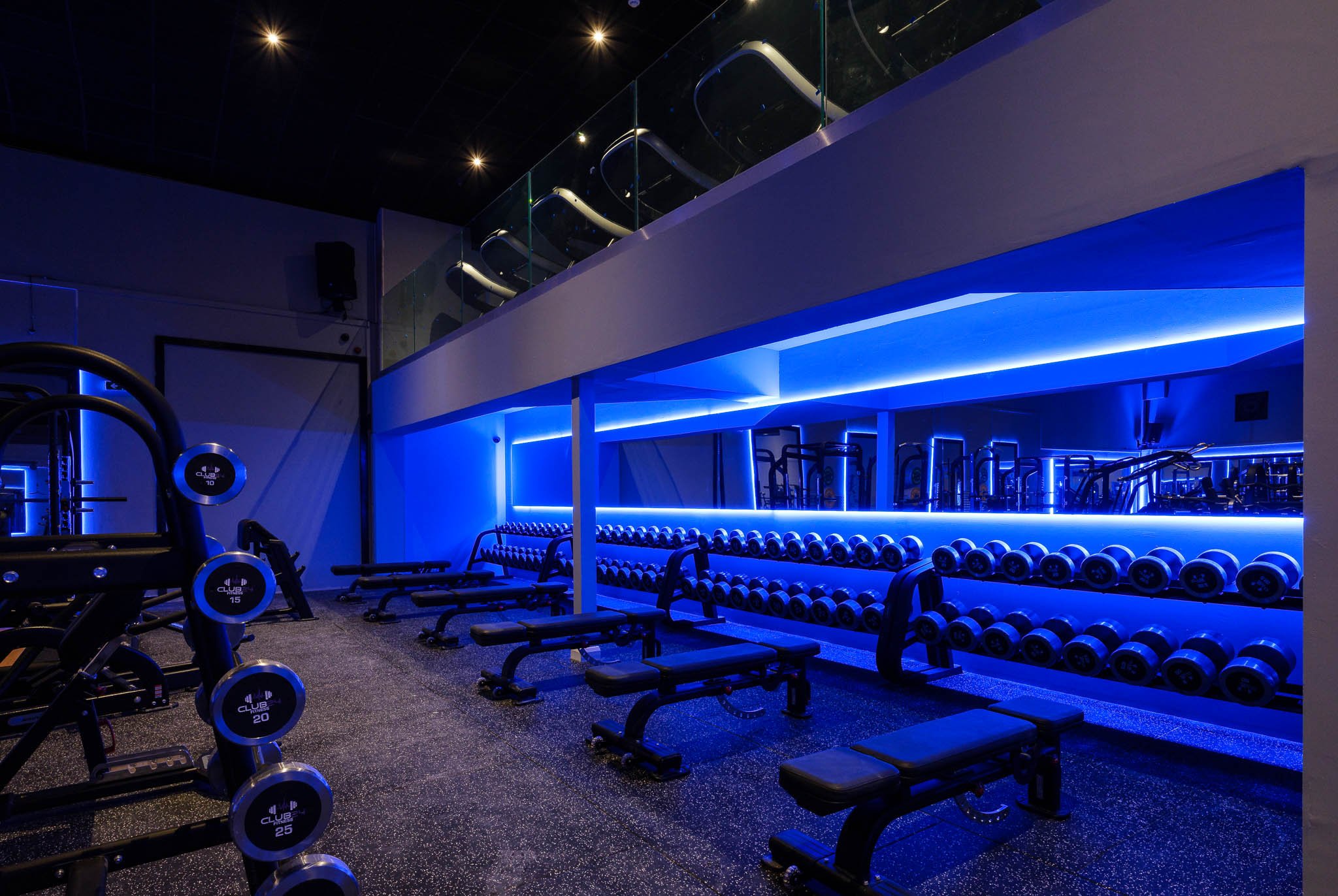 A modern gym with blue LED lighting, including exercise benches, dumbbells on racks, and a second-floor balcony with more workout equipment.