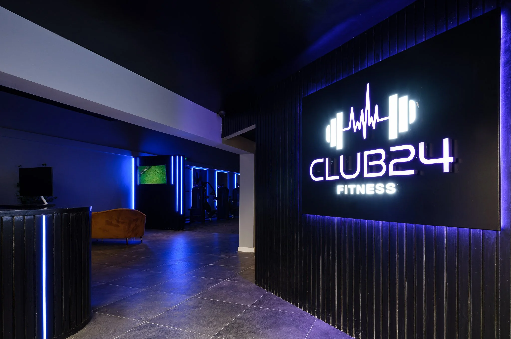 Interior of a fitness club with a prominent illuminated sign reading 'CLUB24 FITNESS' featuring a barbell with a heartbeat line on it. The room has dark walls with blue neon lighting and exercise equipment in the background.