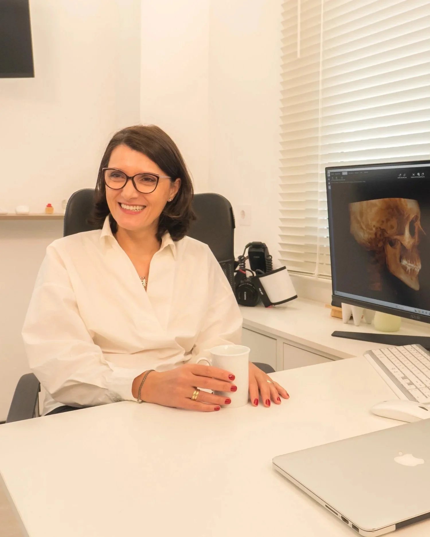 Dentista especializada en ATM y restauraciones complejas en su clínica dental Dra. Violeta Voicu, en el centro de Murcia. Bata blanca, gafas y sonriente, con una pantalla de ordenador que muestra un escaneo de un cráneo para un diagnóstico dental.