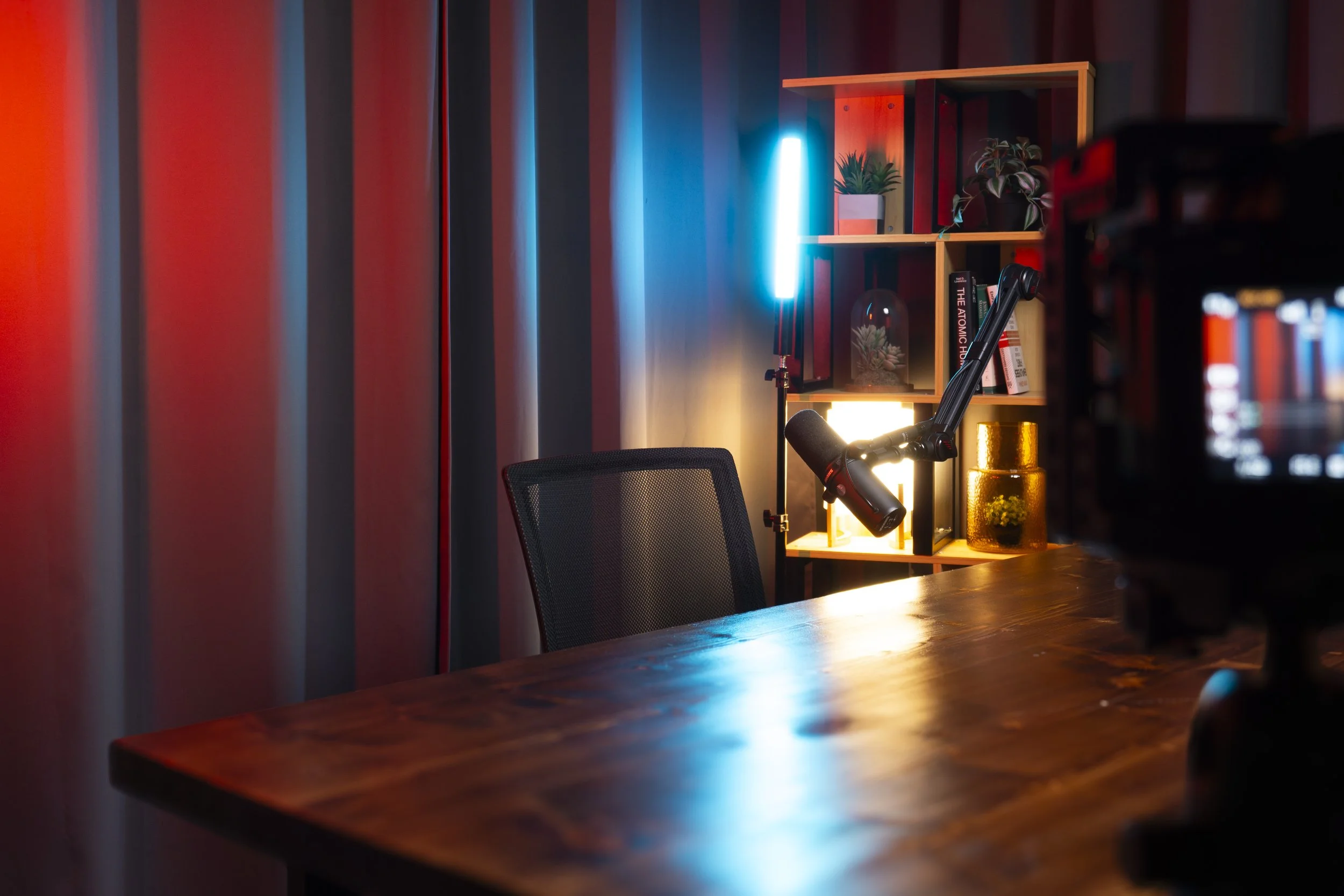 A desk with a microphone and behind it shelves with plants and books, illuminated by colorful LED lights.