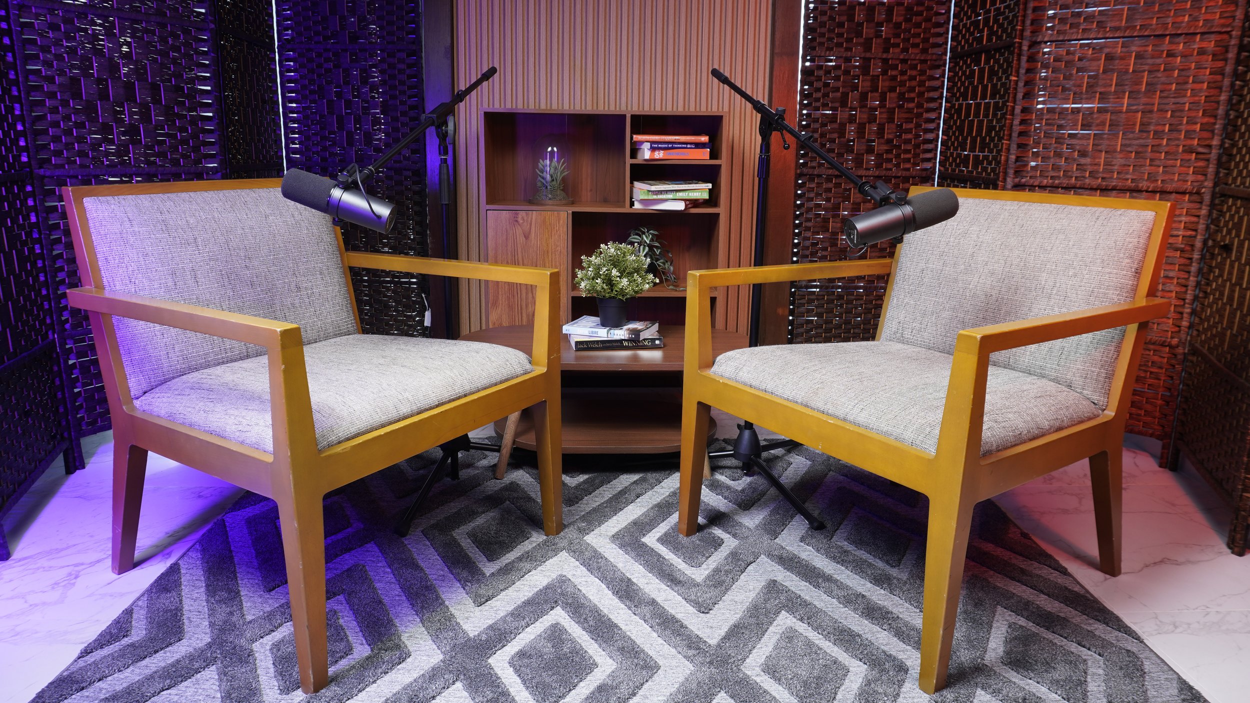Two beige armchairs with wooden frames facing each other with microphones mounted on stands overhead, a wooden bookshelf with plants and books in the background, and a patterned rug on the floor.