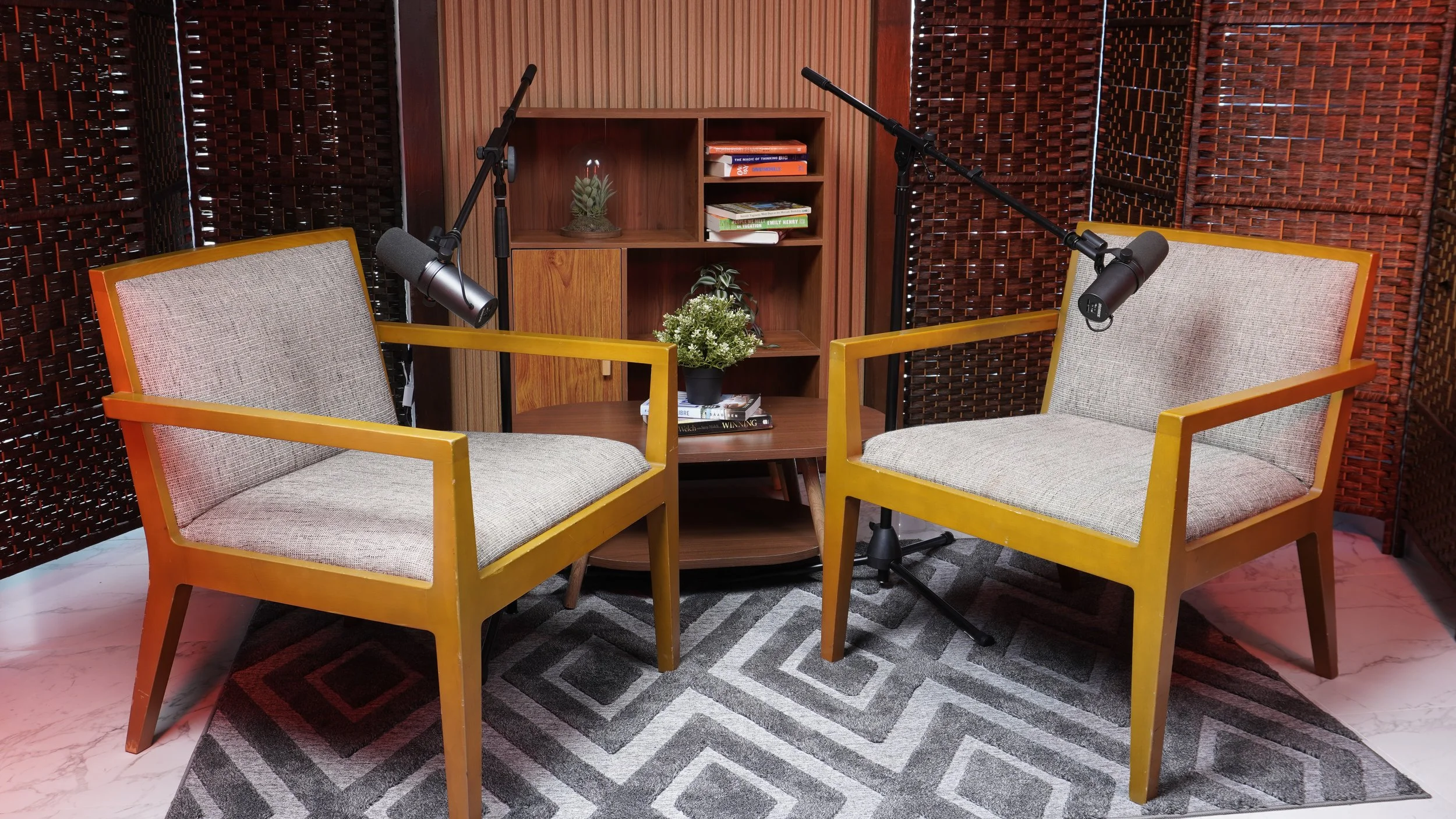 Two beige armchairs with wooden frames facing each other, with studio microphones attached to each armchair, set in a cozy room with wooden panels, a bookshelf, plants, and a patterned rug.