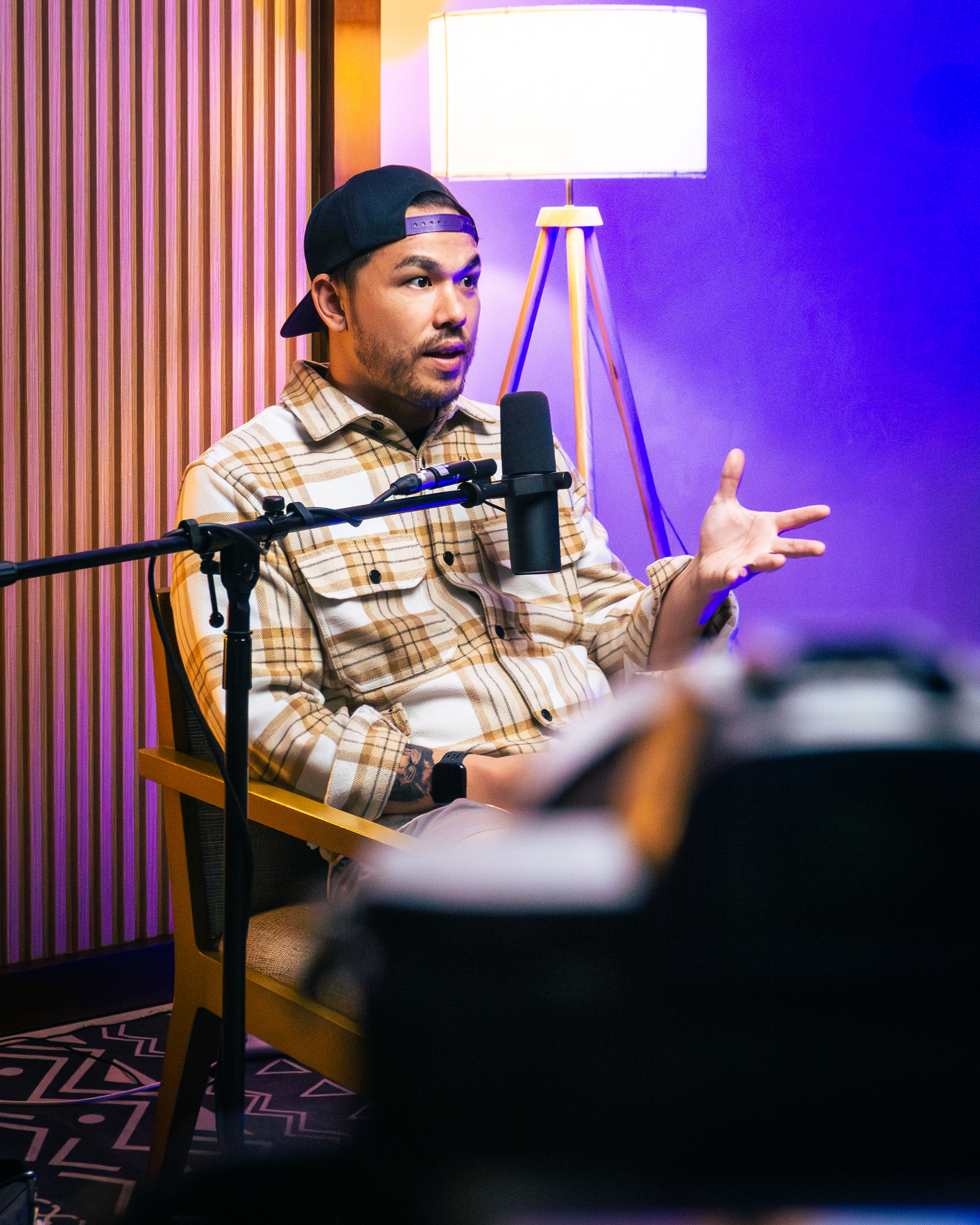 A man wearing a black cap backwards and a checkered shirt is sitting in a studio, speaking into a microphone. He is gesturing with his right hand, and a lamp with a wooden tripod base is behind him, casting warm light. The studio has purple and yellow lighting, with wooden paneling on one wall and a patterned carpet.