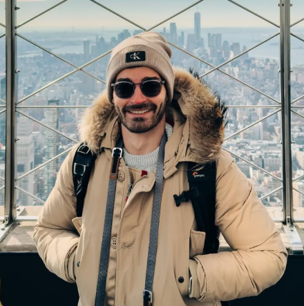 Smiling man wearing a beige winter coat, beanie, and sunglasses, standing on a high observation deck with a city skyline in the background.