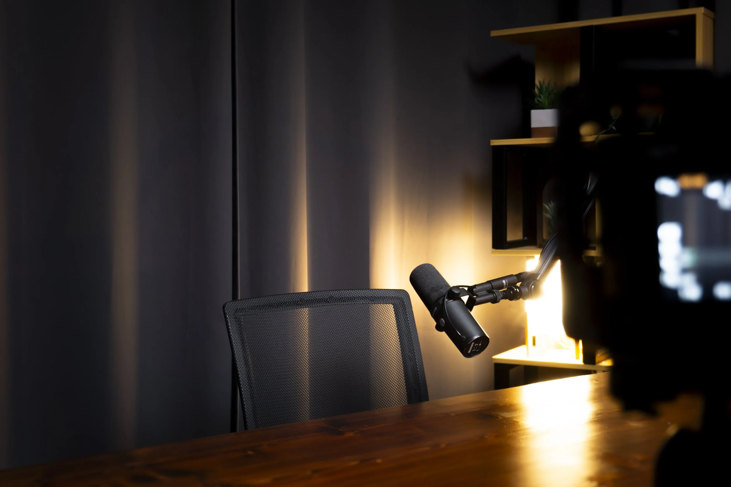 A recording studio setup with a black microphone mounted on an adjustable arm, a black office chair, and a warm light source illuminating a dark wall and shelves with decorative items and plants.