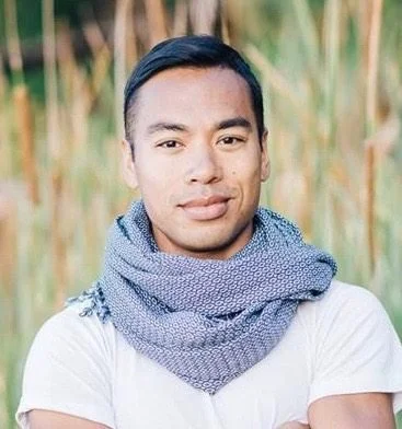 Portrait of a young man with short black hair, wearing a white T-shirt and a blue scarf, standing outdoors with tall grass or reeds in the background.