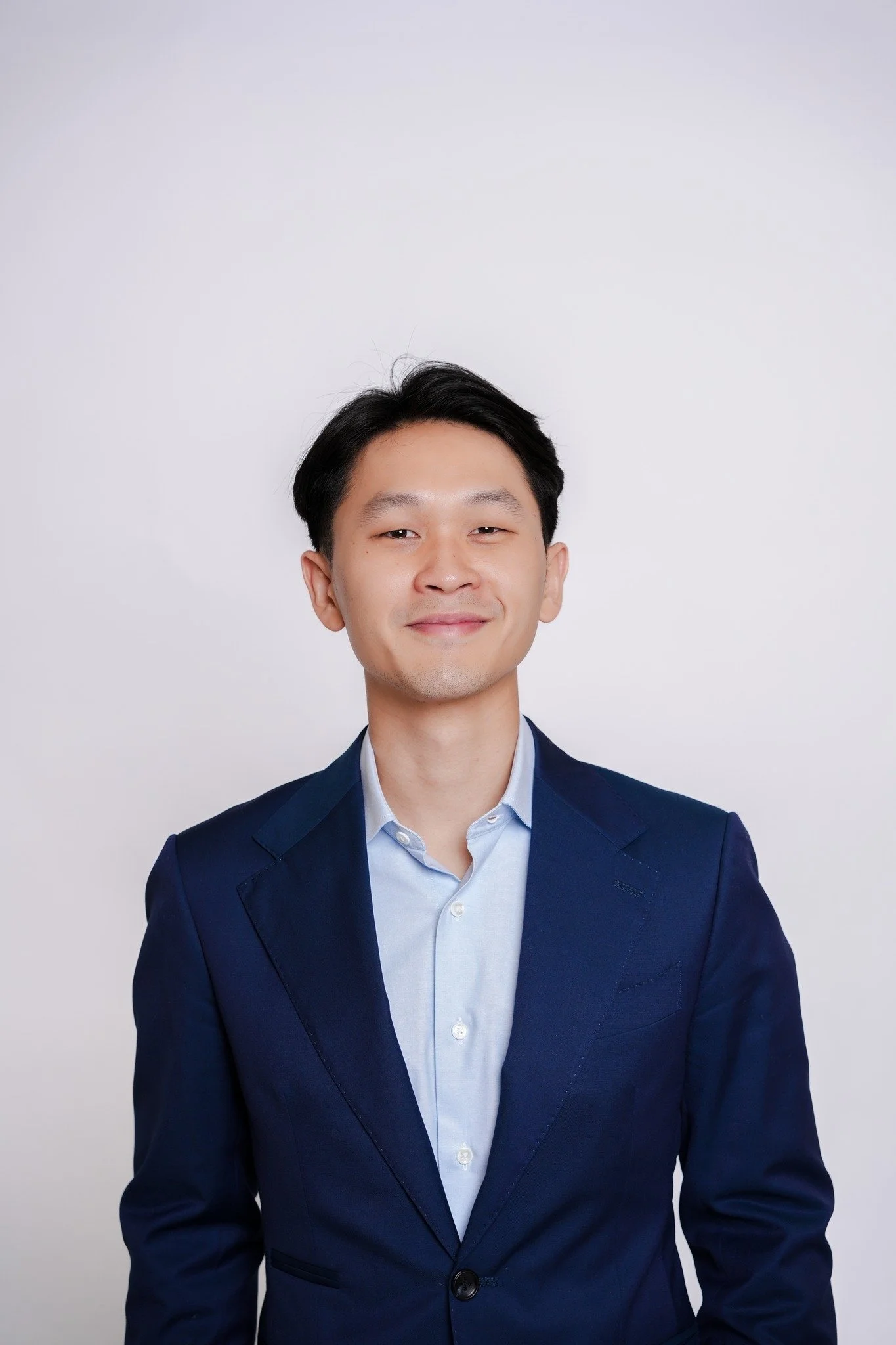 A young man with black hair wearing a dark blue suit and light blue shirt, standing against a plain white background.