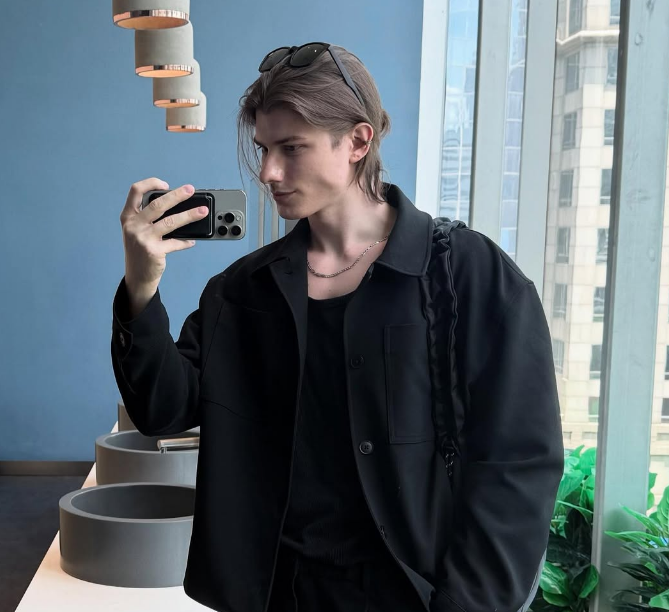 Young man taking a mirror selfie in a modern room with blue walls and large windows, wearing sunglasses on his head, black jacket, and holding a smartphone.