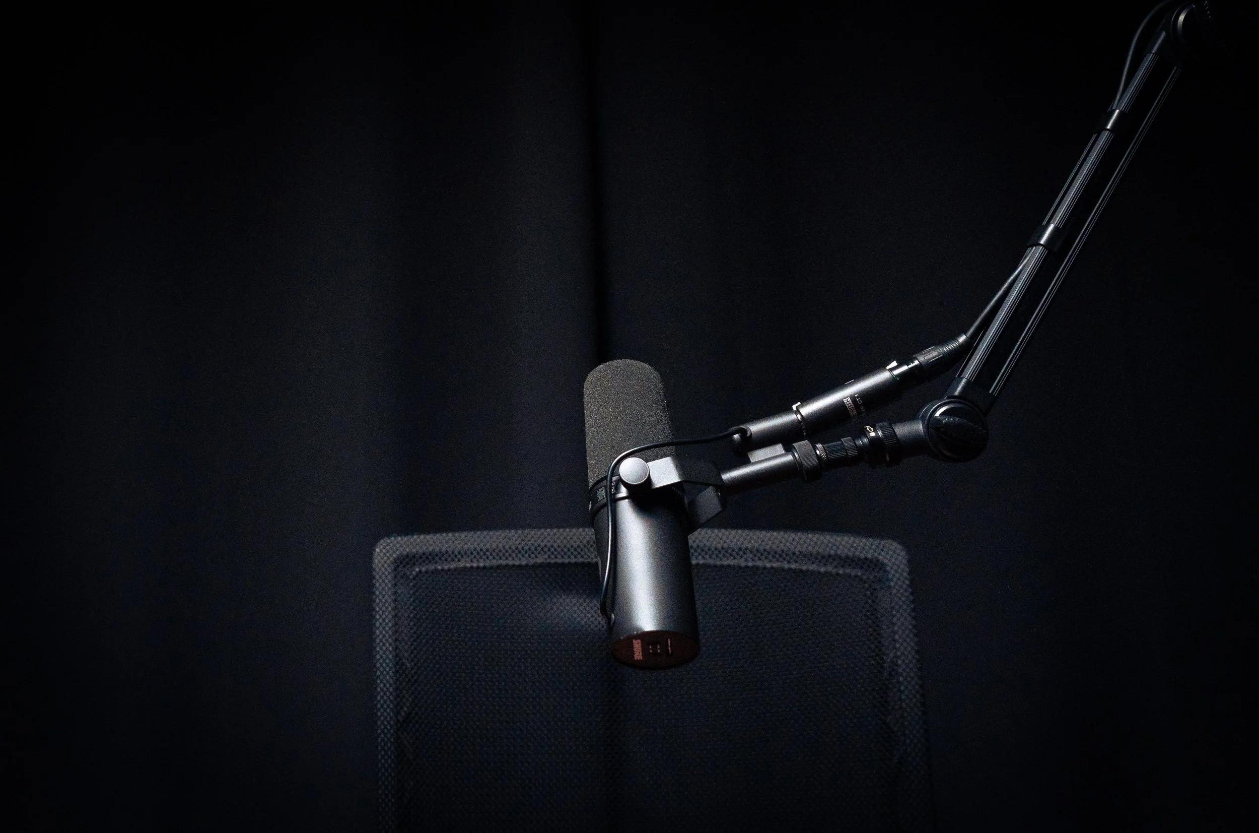 A professional microphone mounted on an adjustable boom arm in a sound recording studio with a dark background.