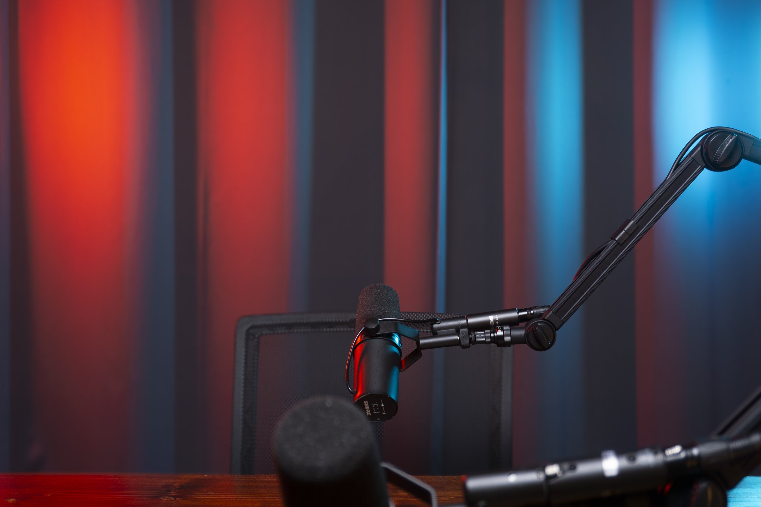 A professional recording studio setup with a microphone on a boom arm, a podcasting microphone, and an office chair, against a background of vertically striped red and blue curtains.