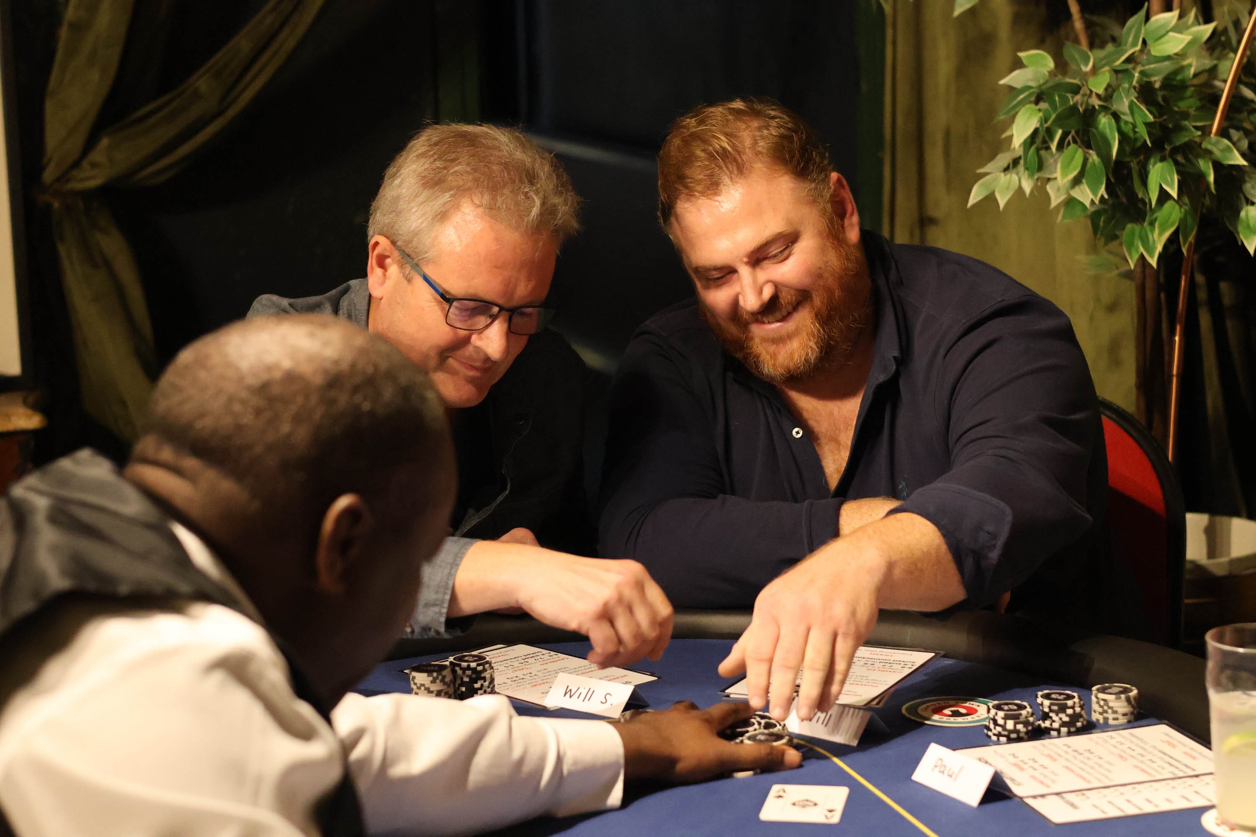 Three men playing poker at a table, smiling and engaging while playing Texas Hold'em poker chips, with playing cards and handout instruction or poker tutorials visible. There is a poker tutor, tutorial or professional dealer visible.