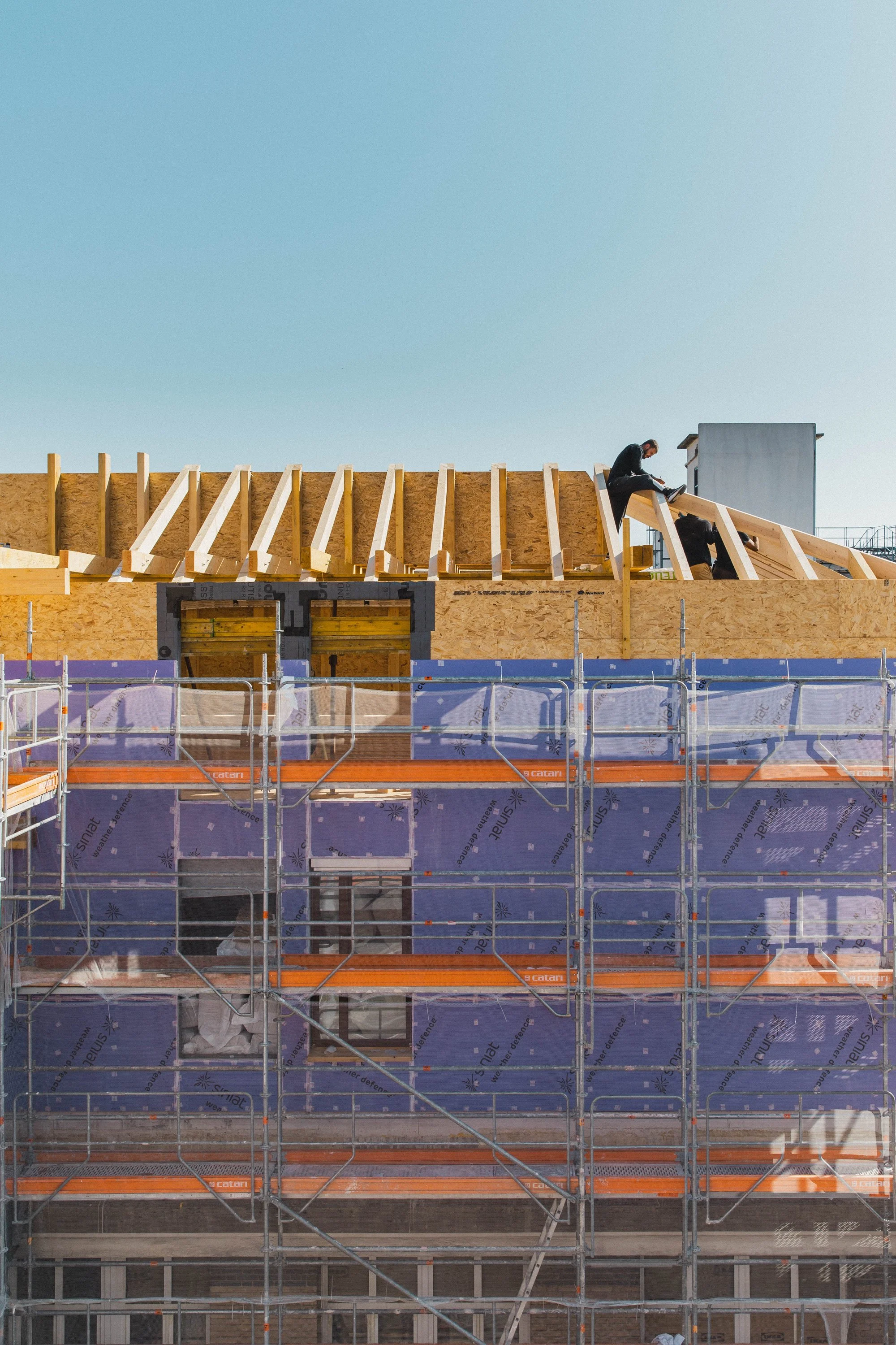 Travaux de construction sur un bâtiment avec des ouvriers posant la toiture, des échafaudages et une façade recouverte d'une aislant.