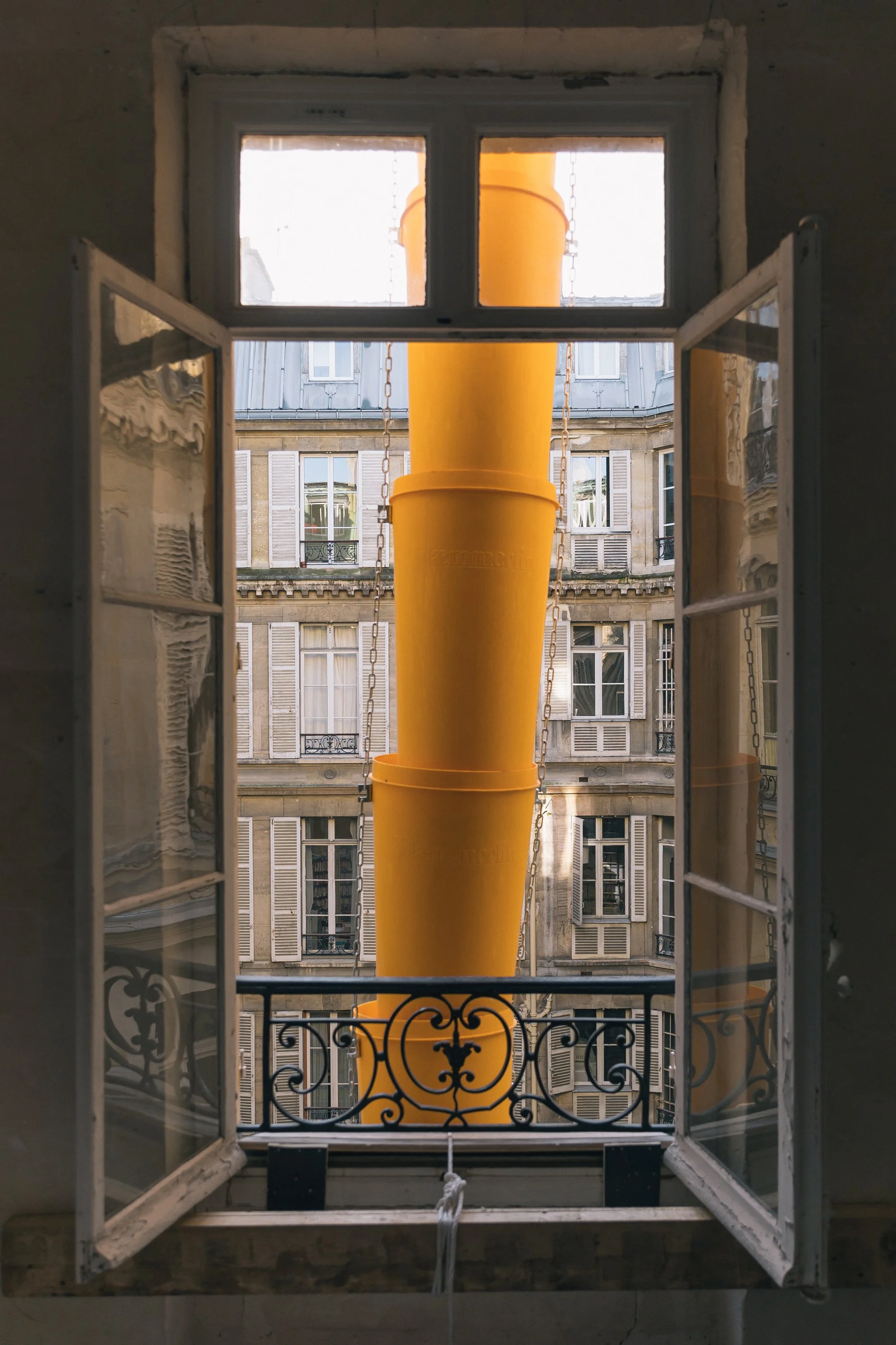Fenêtre ouverte avec vue sur un bâtiment haussmannien et un tuyau de chantier jaune suspendu en dehors.
