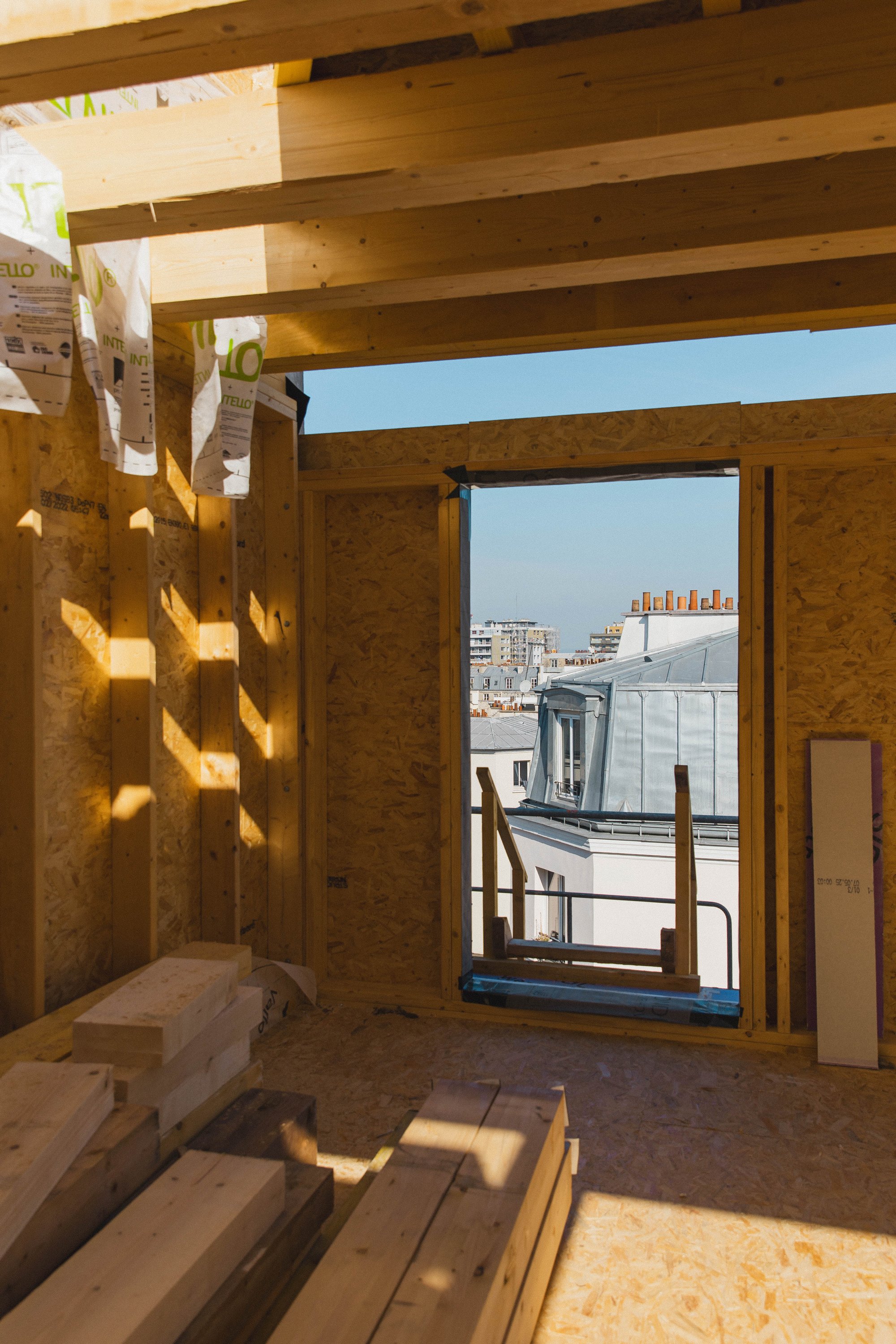 Intérieur de la construction d'un bâtiment avec murs en bois, fenêtre ouverte et vue sur des bâtiments en ville lors d'une journée ensoleillée.