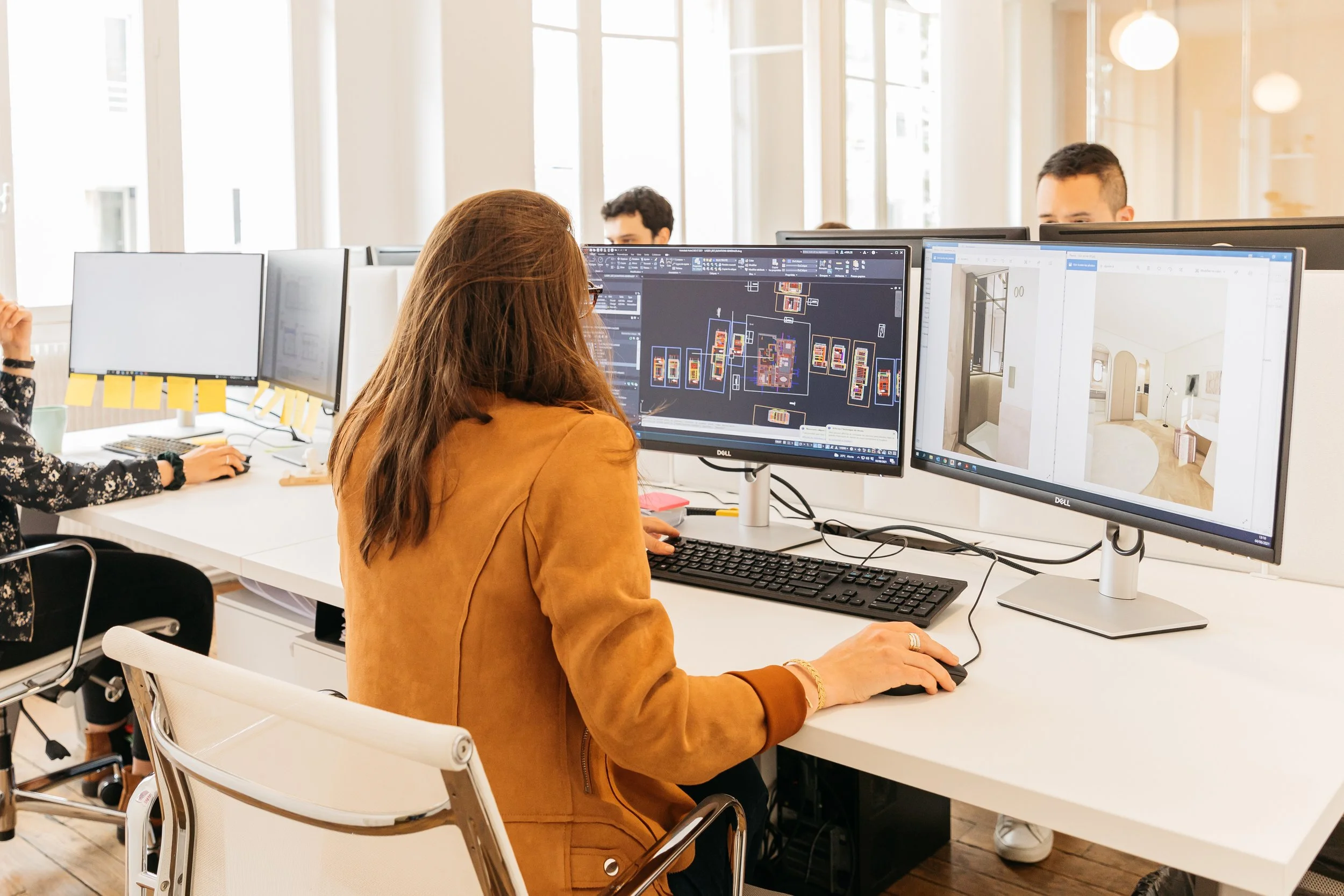 Une femme travaille sur deux écrans d'ordinateur, dont l'un affiche un plan architectural et l'autre une conception intérieure, dans un bureau lumineux avec de grandes fenêtres.