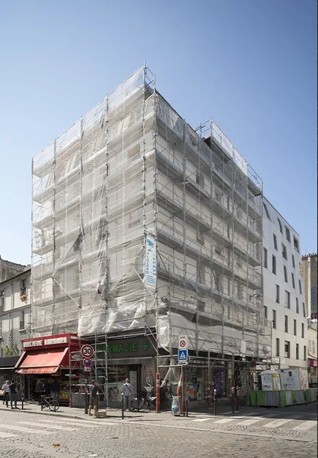 Bâtiment en rénovation avec échafaudages recouverts de bâches, situé à un coin de rue avec des passants et des commerces en dessous.
