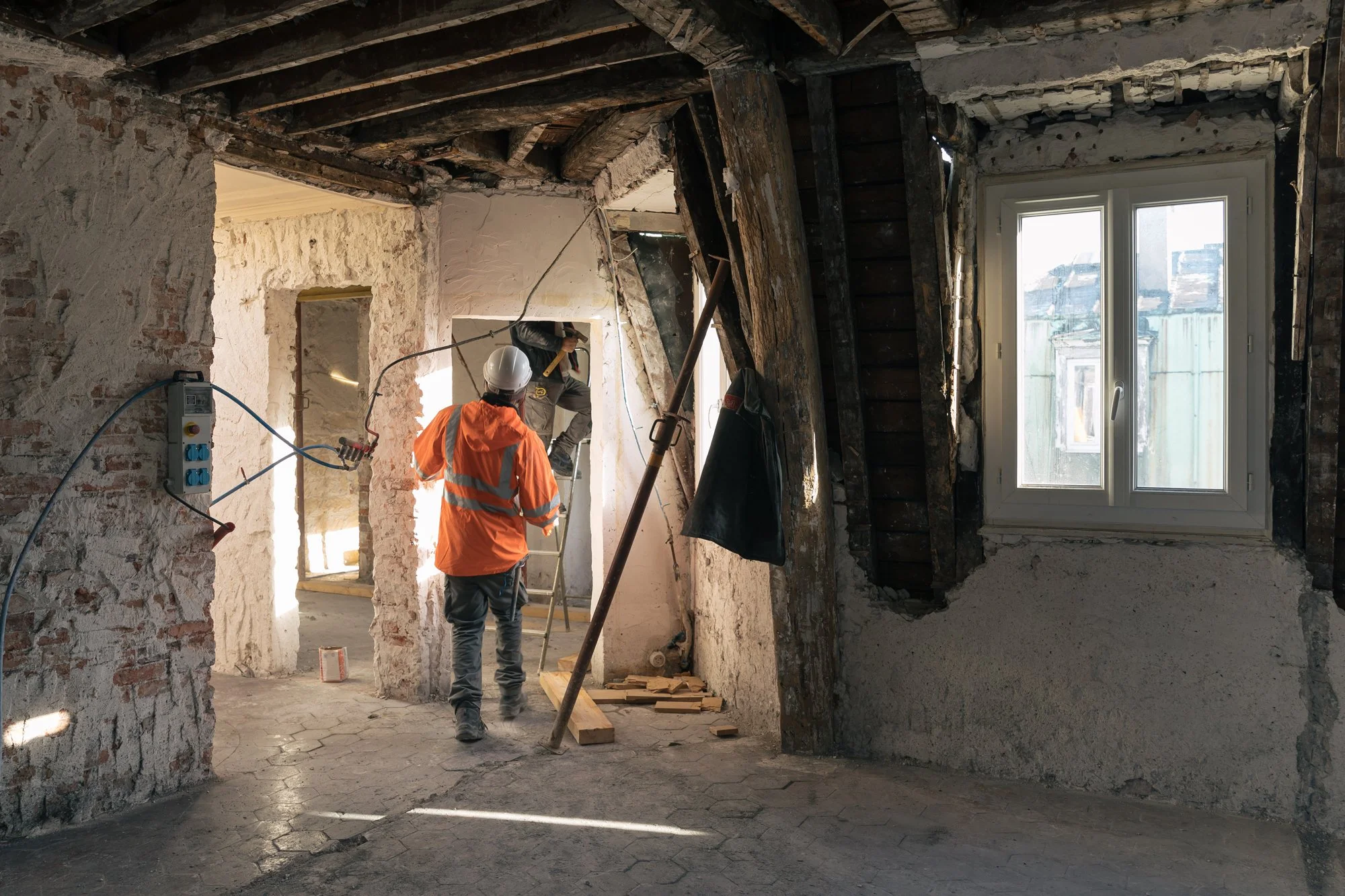 Travaux de rénovation intérieure avec deux ouvriers portant des casques de sécurité dans un bâtiment ancien avec murs en pierre et une fenêtre.