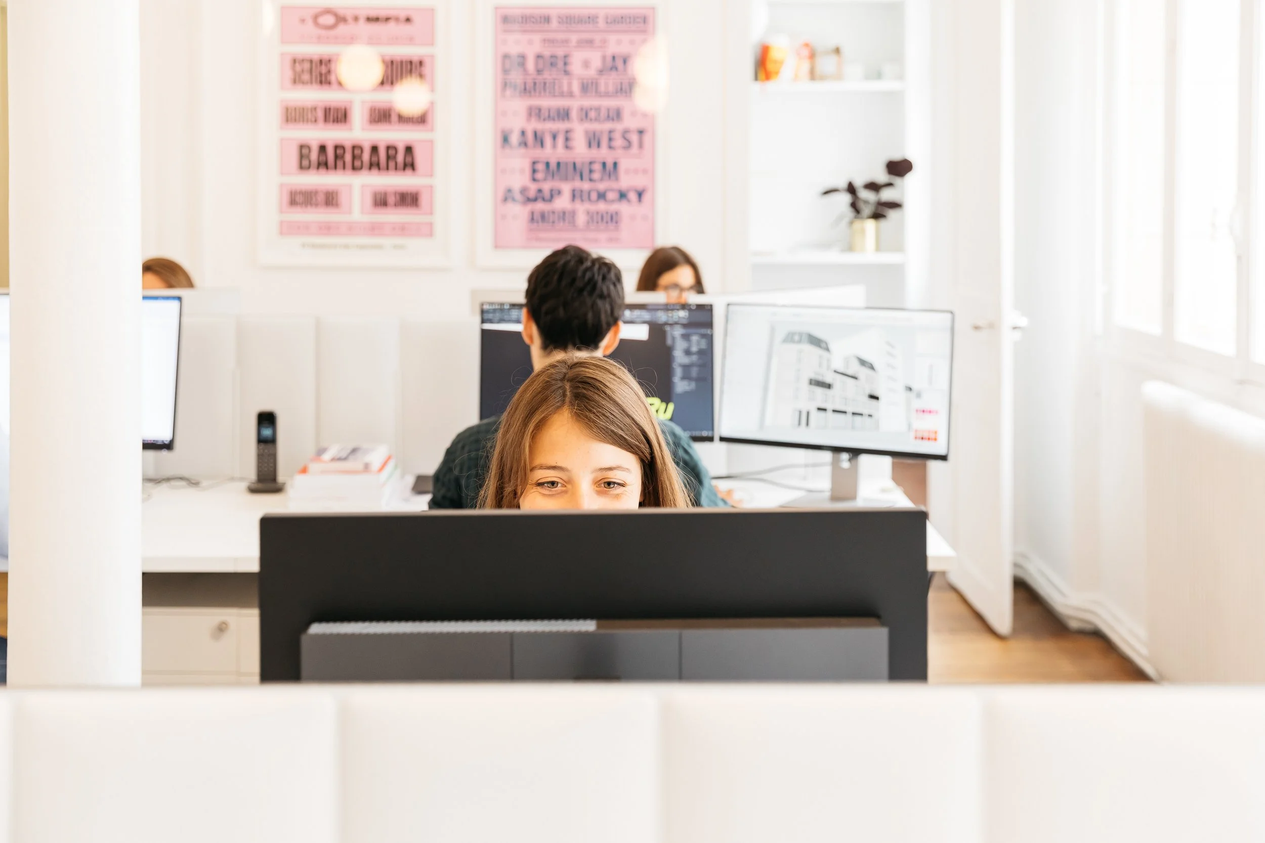 Groupe de personnes travaillant dans un bureau avec plusieurs écrans d'ordinateur, posters roses sur le mur et fenêtre donnant sur l'extérieur.
