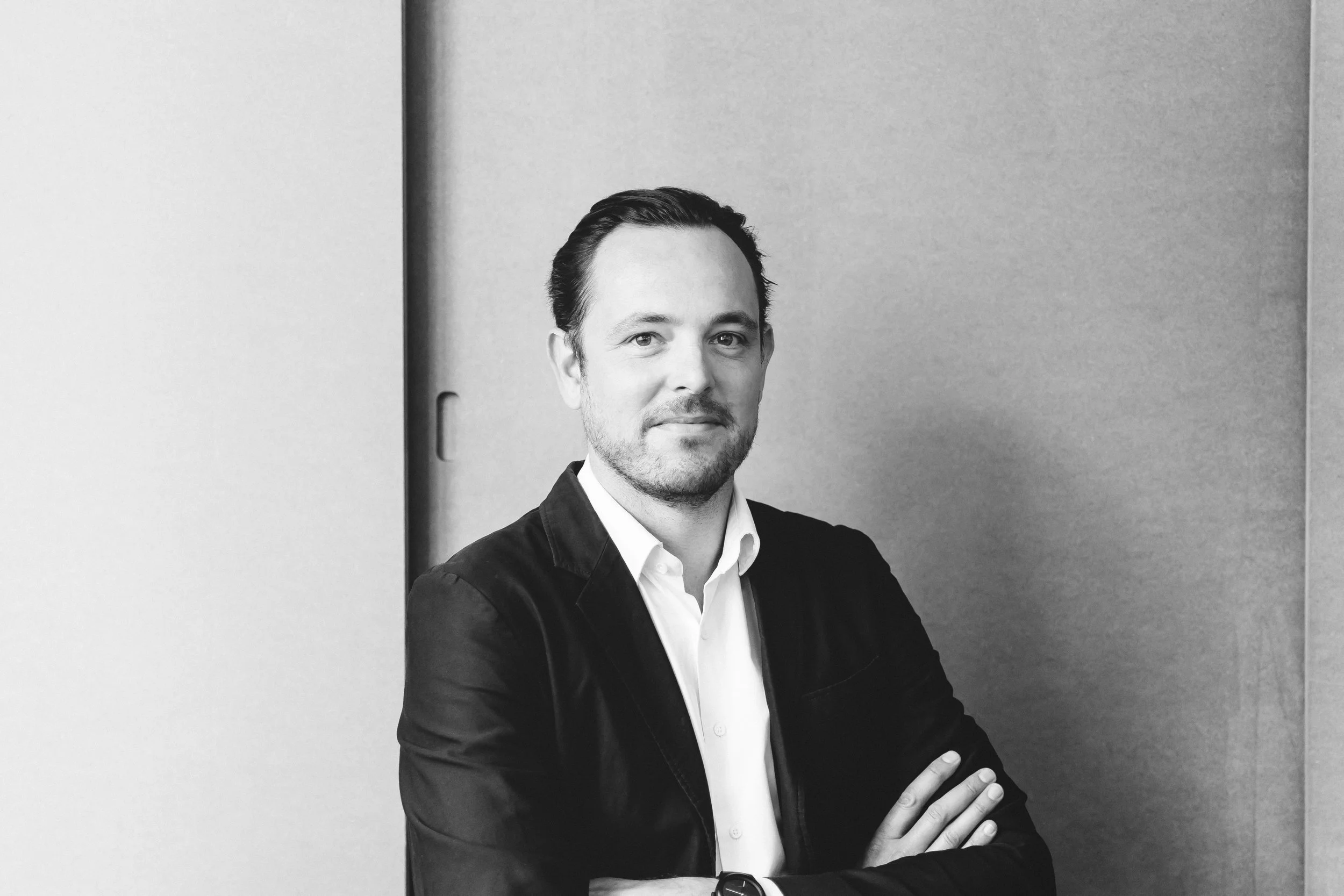 Portrait en noir et blanc d’un homme d’apparence professionnelle, portant une chemise blanche et une veste sombre, avec les bras croisés, dans un environnement d’intérieur.