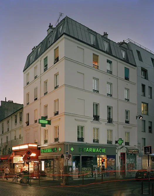 Bâtiment avec pharmacie au rez-de-chaussée, en soirée, avec plusieurs étages et un toit en zinc, window lighting visible dans certains appartements.