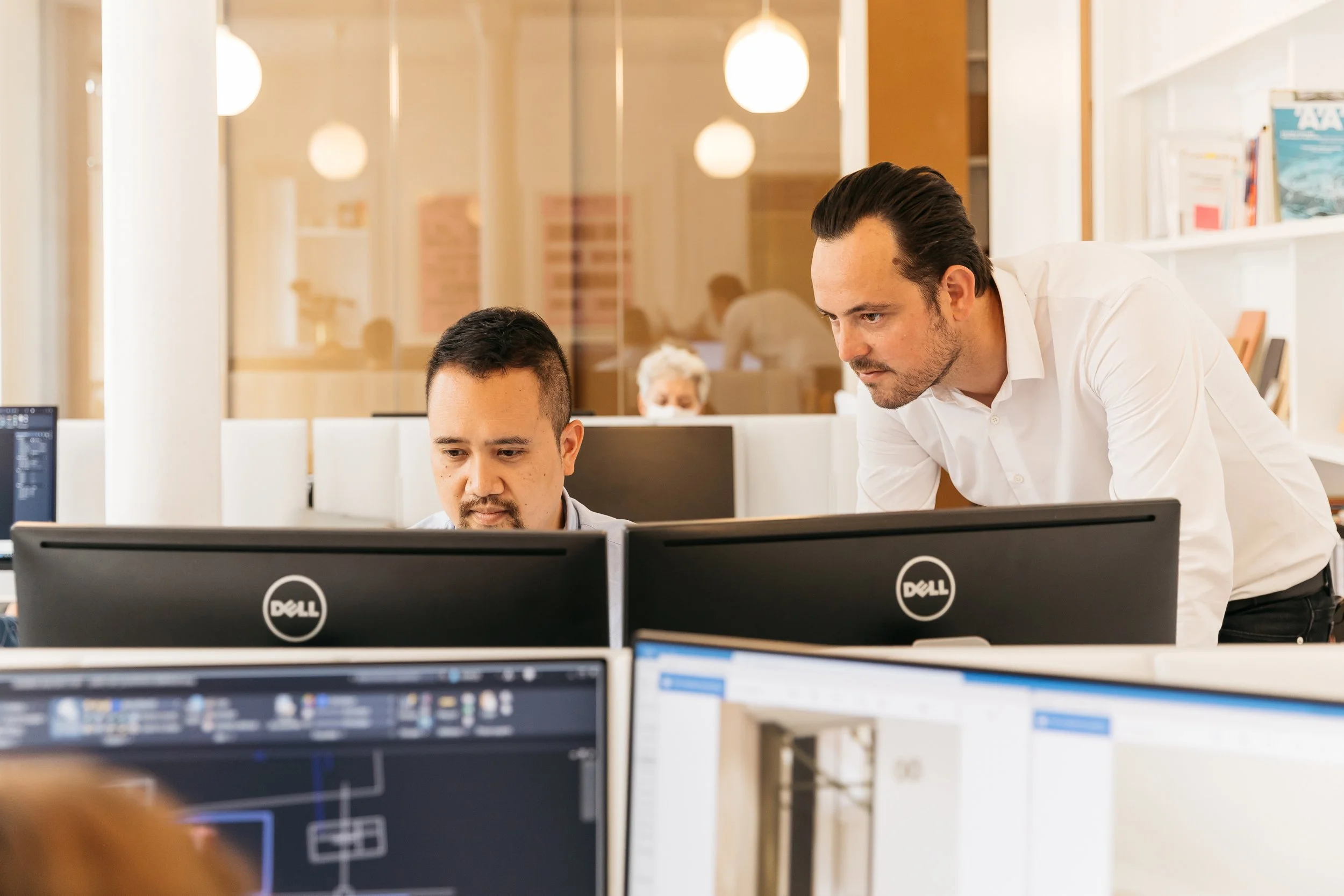 Deux hommes au bureau, l'un regardant l'écran d'ordinateur de l'autre, en mode de collaboration, dans un environnement de travail moderne.