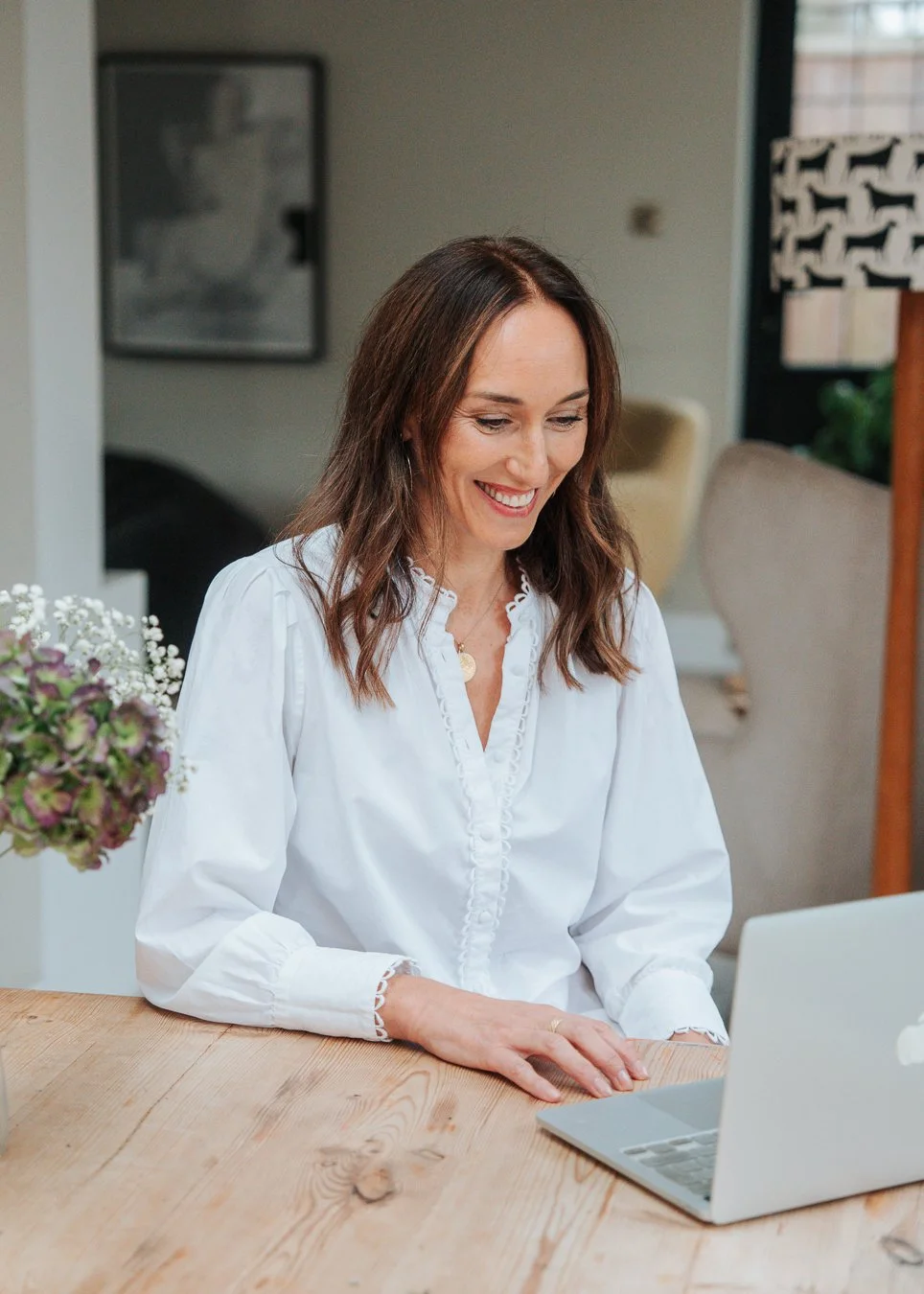 A woman with shoulder-length brown hair wearing a white blouse, sitting at a wooden table, smiling while looking at a silver laptop.