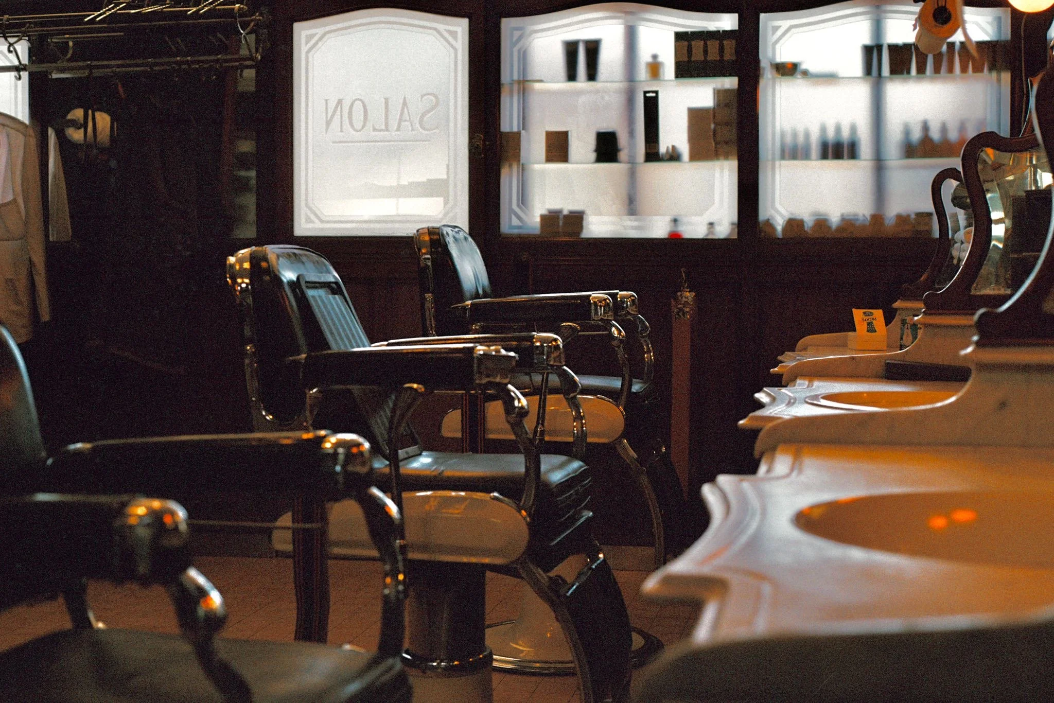 Interior of a vintage barbershop with chairs in front of sinks, dark wooden paneling, and large frosted windows.