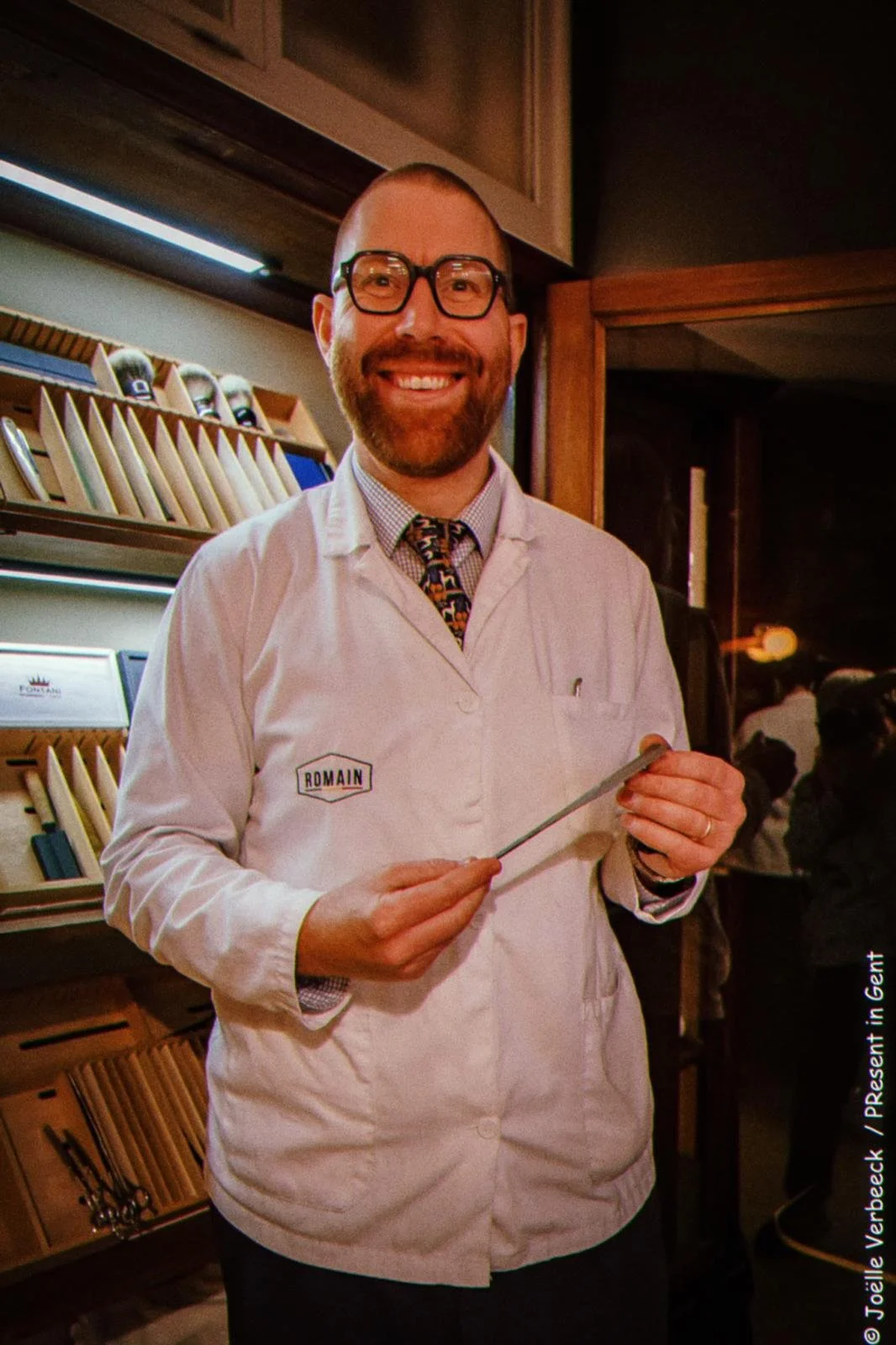 A smiling man with glasses and a beard wearing a white lab coat holding a metal instrument, standing indoors with a background of cutting boards and other kitchen items.