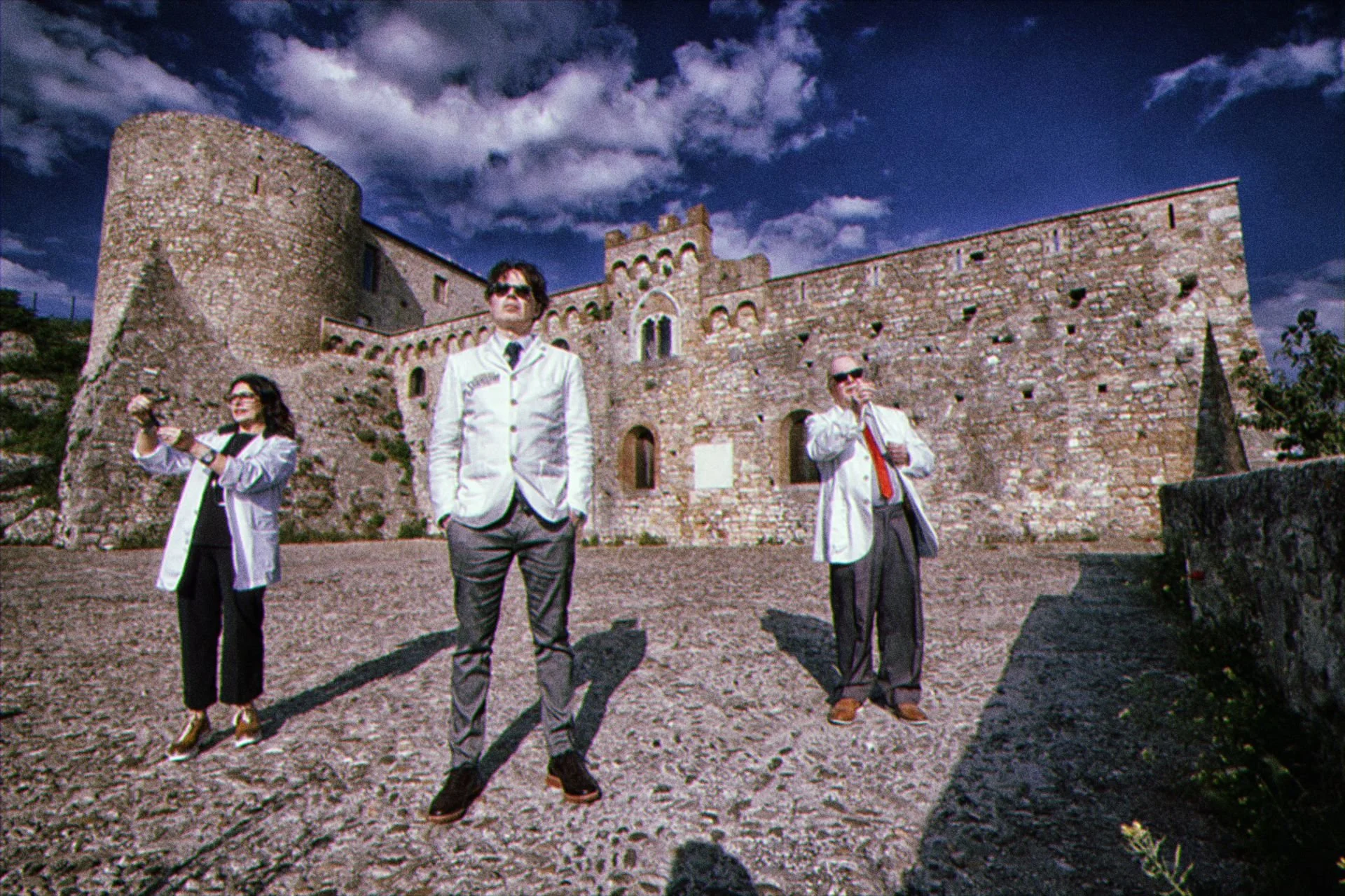 Three people in formal attire, standing in front of an old stone castle under a cloudy sky.