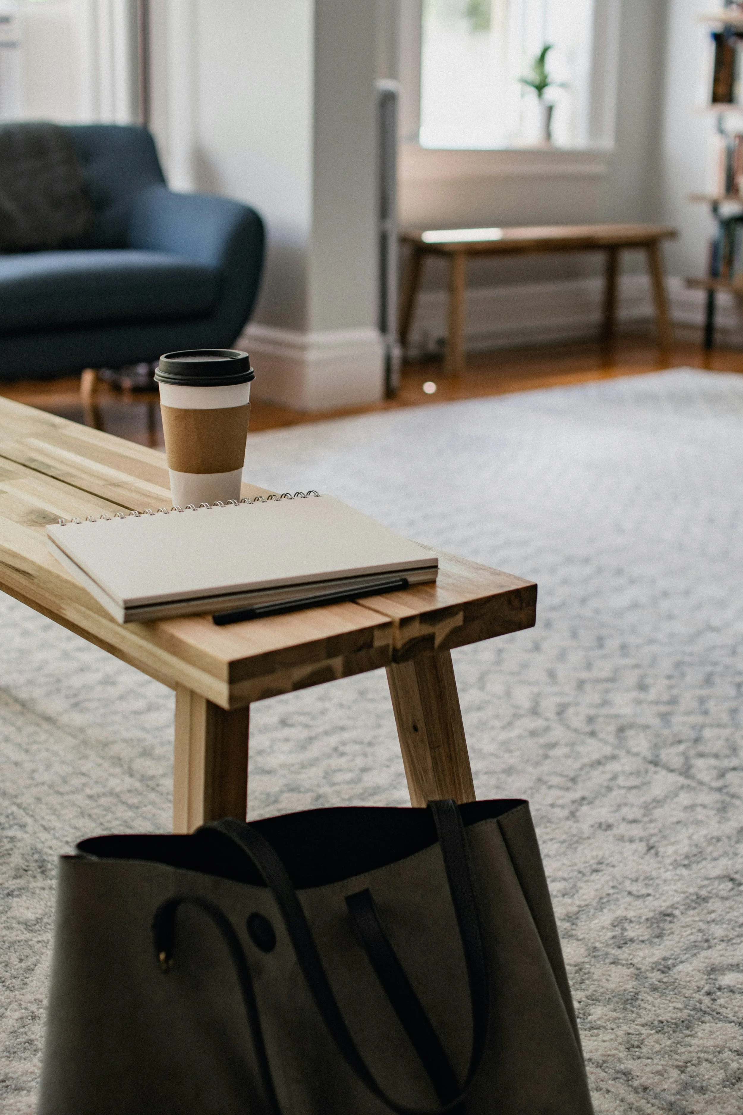 A wooden table holds a spiral notebook, a black pen, and a coffee cup. In the foreground, a black and gray tote bag is visible. The background shows a living room with a dark blue couch, a window, a small wooden bench, a white rug, and bookshelves.