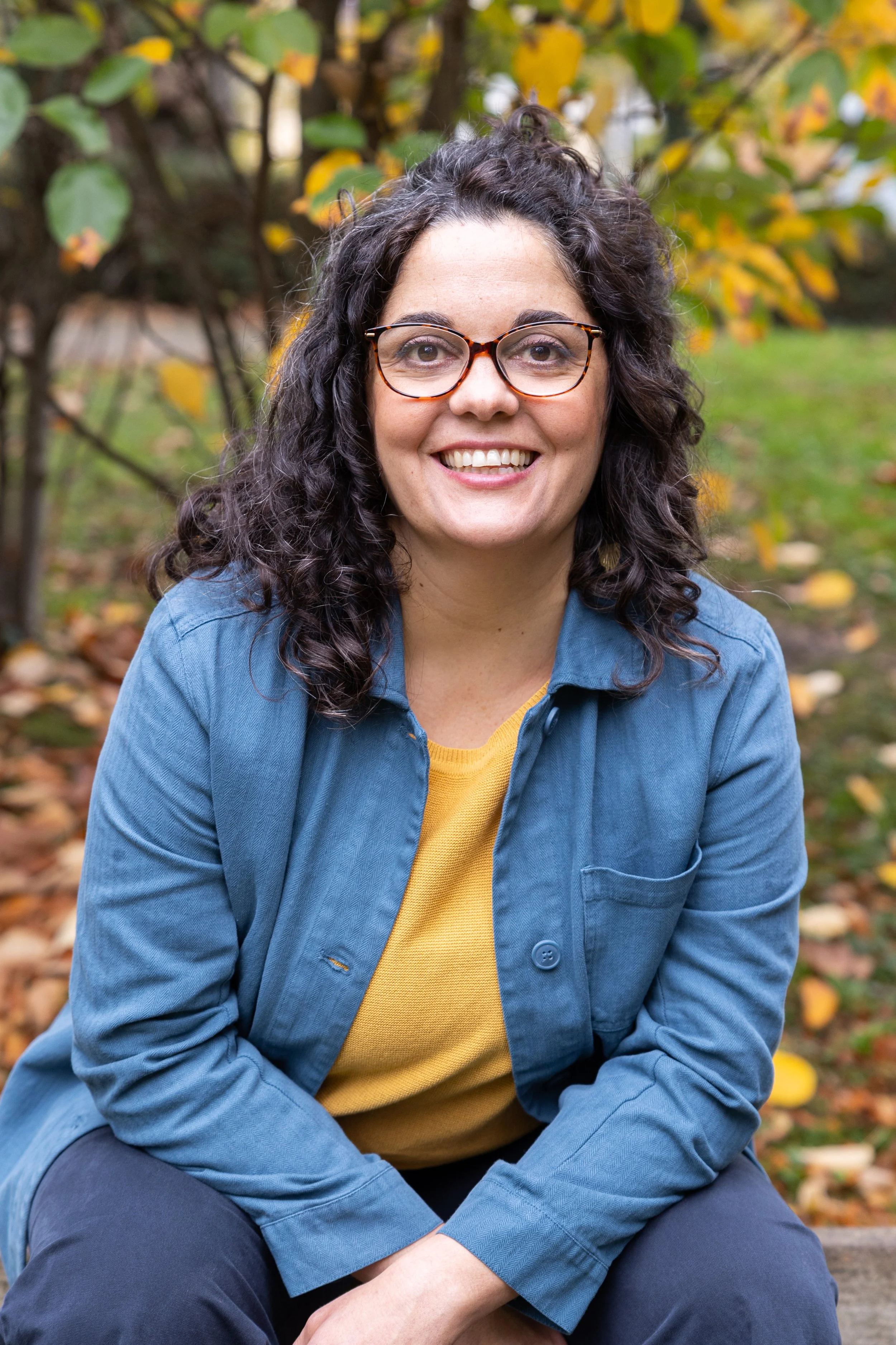 A woman with curly dark hair, wearing glasses, a blue jacket, and a yellow shirt, smiling outdoors with autumn leaves in the background.
