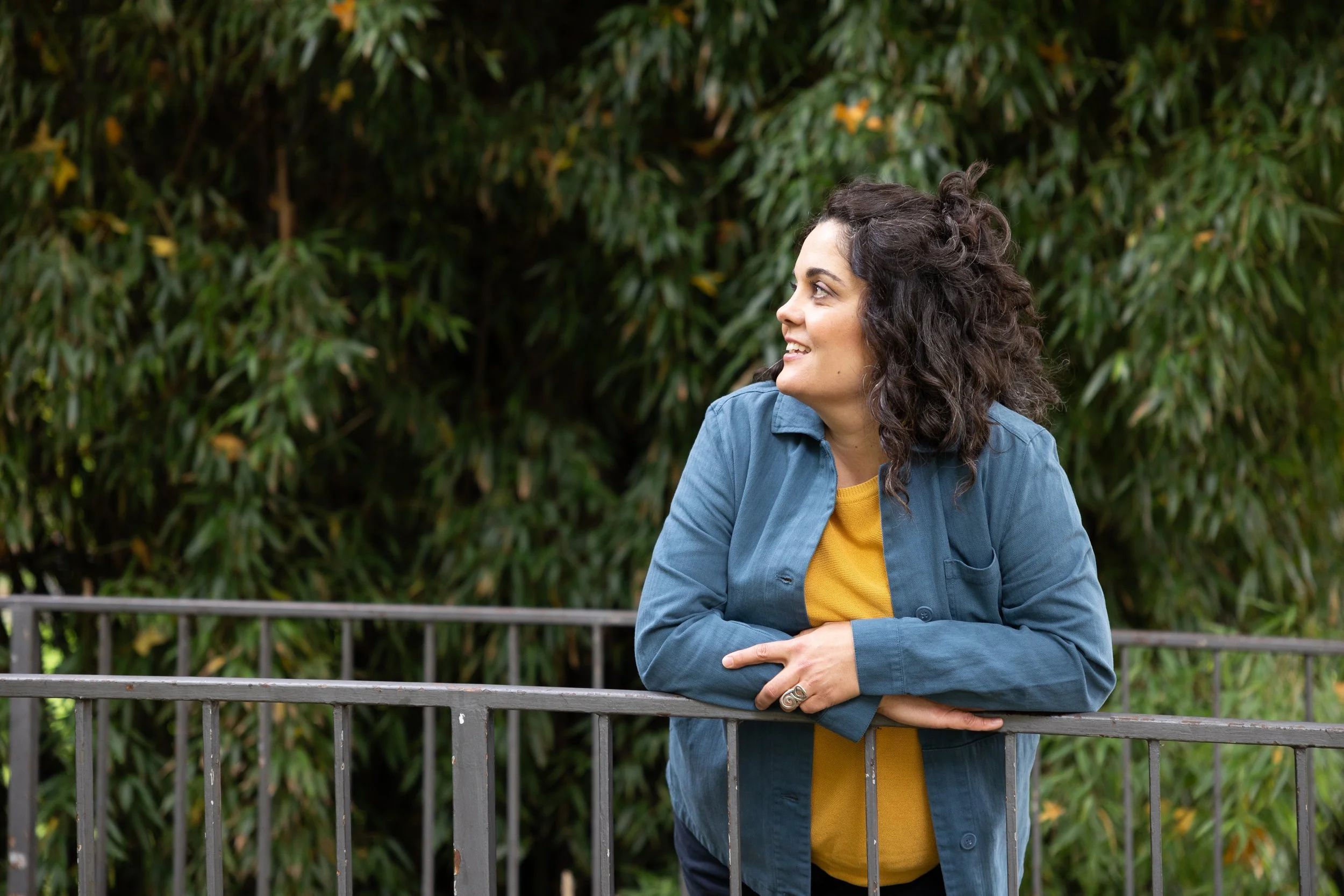 A woman with curly dark hair, wearing a yellow shirt and a blue jacket, leaning on a metal railing outdoors with green foliage in the background, smiling and looking to the side.