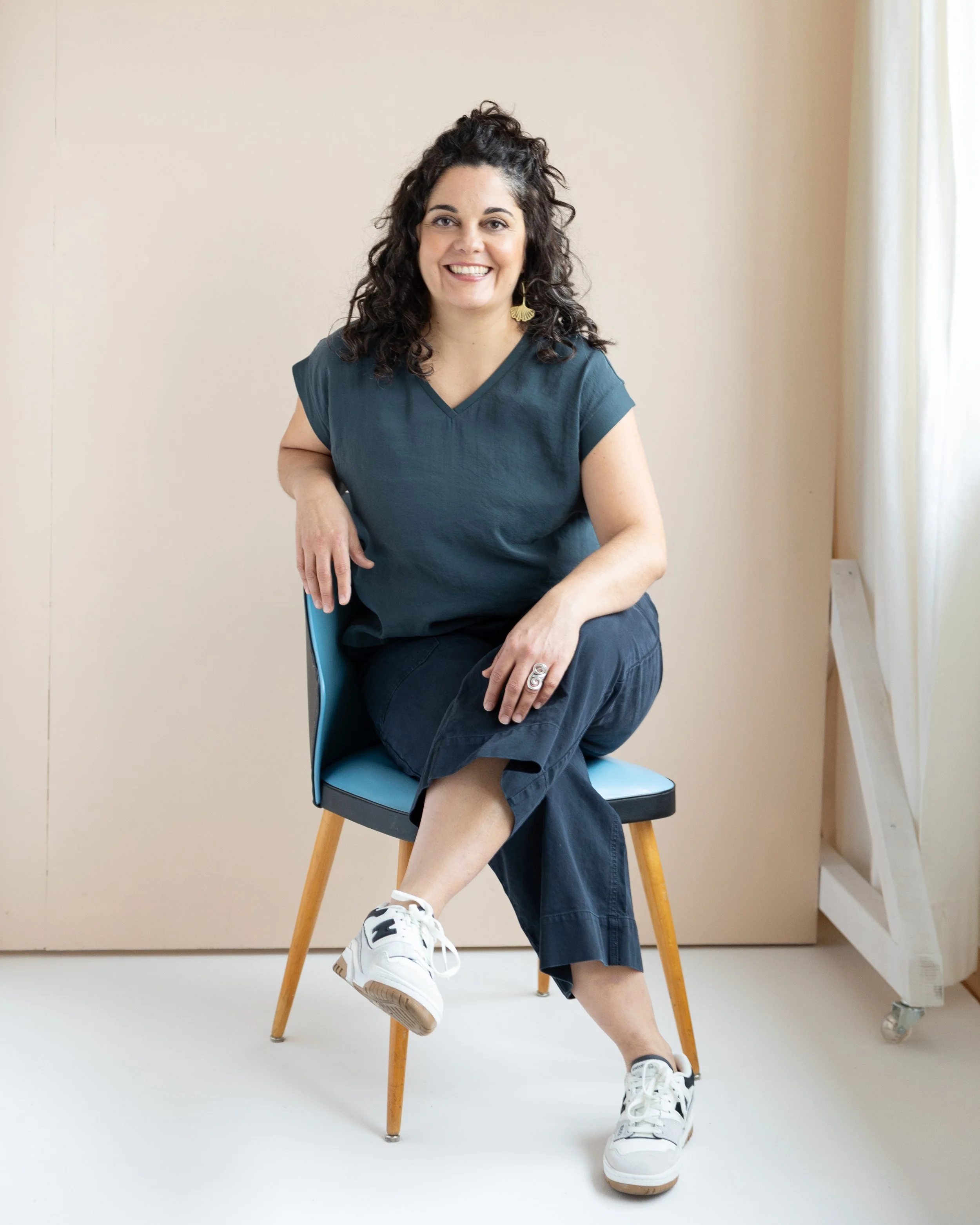 A woman with curly dark hair, smiling, sitting on a blue chair with wooden legs, wearing a teal top, black pants, and white sneakers, in a room with light-colored walls and curtains.
