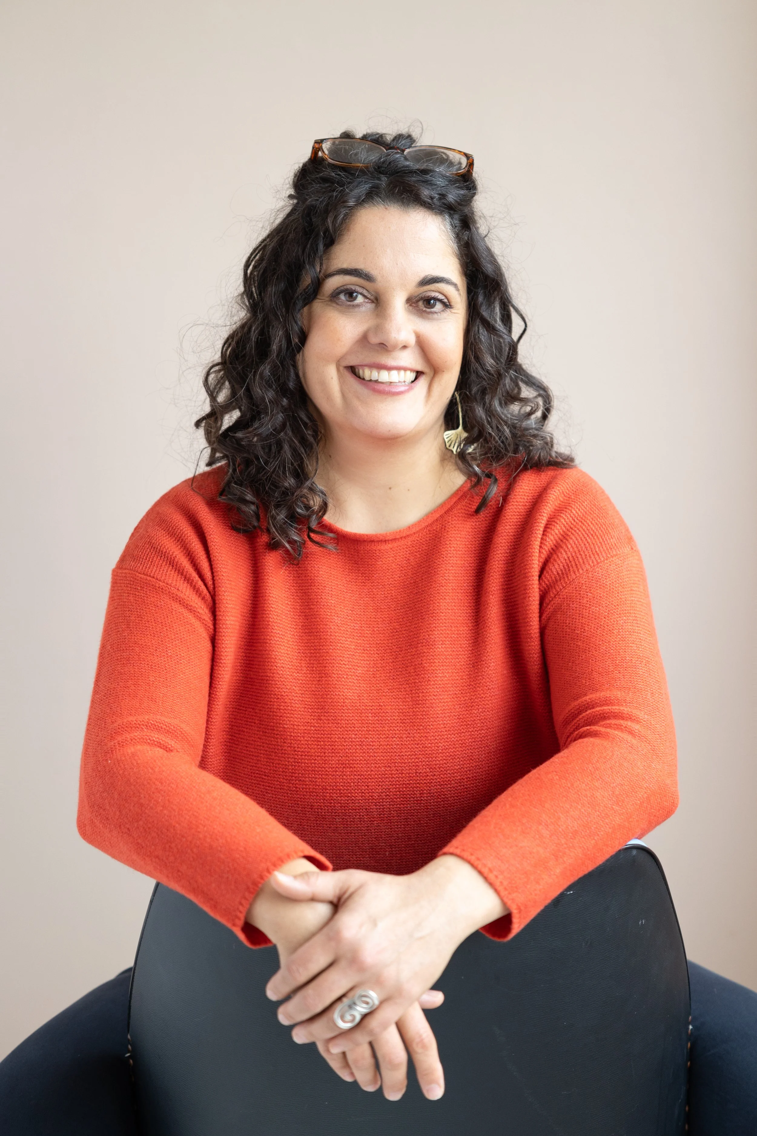 A woman with dark, curly hair and glasses on her head, smiling, wearing an orange sweater, sitting on a black chair with a plain beige background.