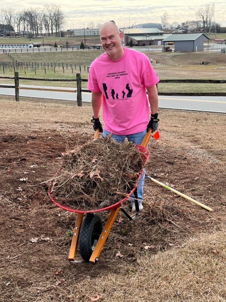 A man smiling while standing outdoors, holding a wheelbarrow filled with dirt and branches, wearing a bright pink T-shirt and gloves.