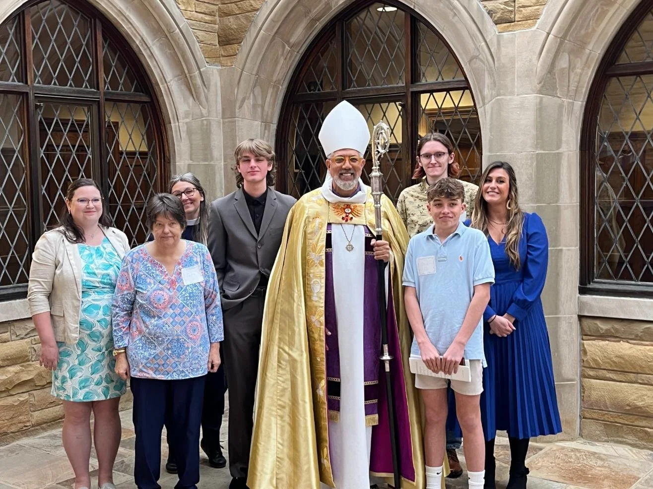 Group of nine people, including a bishop in religious vestments, standing inside a stone building with large arched windows.