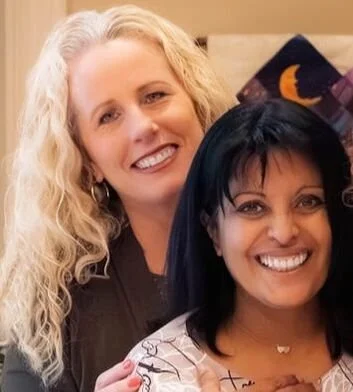 Two women smiling, one with blonde curly hair and the other with straight black hair, in a warmly lit room.