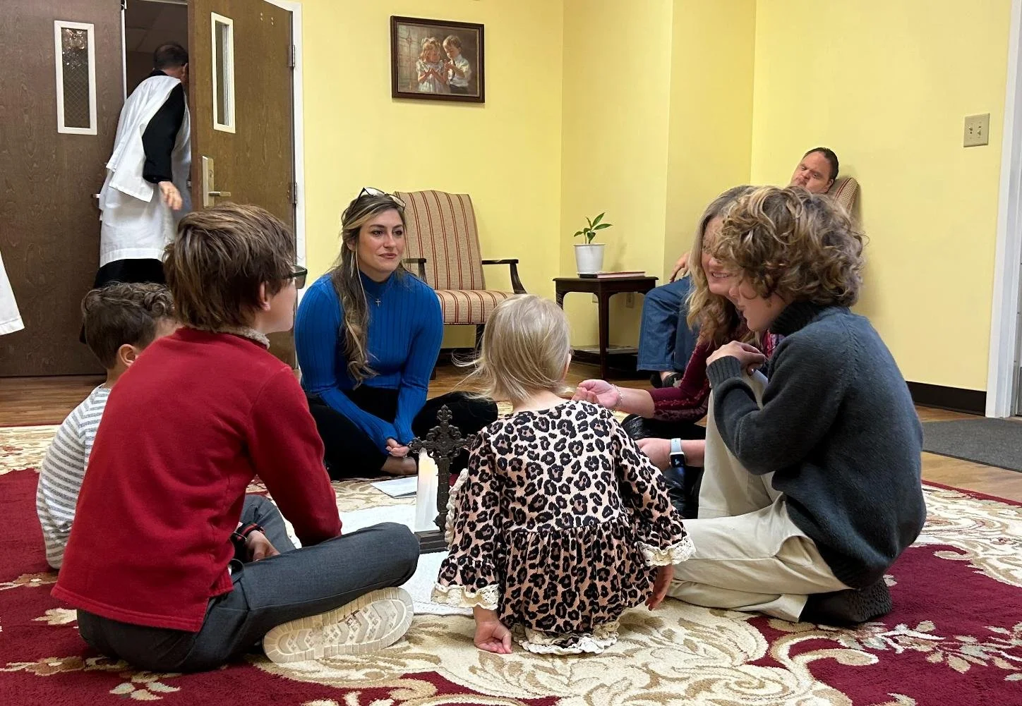 Group of people sitting on a carpet indoors, engaged in conversation, with a woman approaching from the doorway in the background.